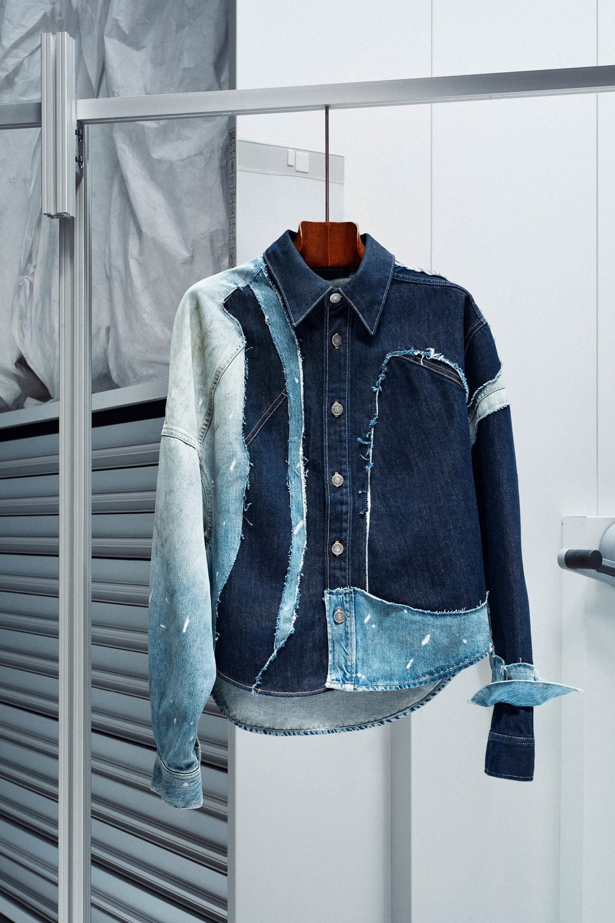 The Acne Studios Hussein Chalayan Patchwork Denim Shirt on a wooden hanger in an archive store room: dark rinse denim pieced with raw-edged panels of pale, bleach-spotted denim down one side and across the hem