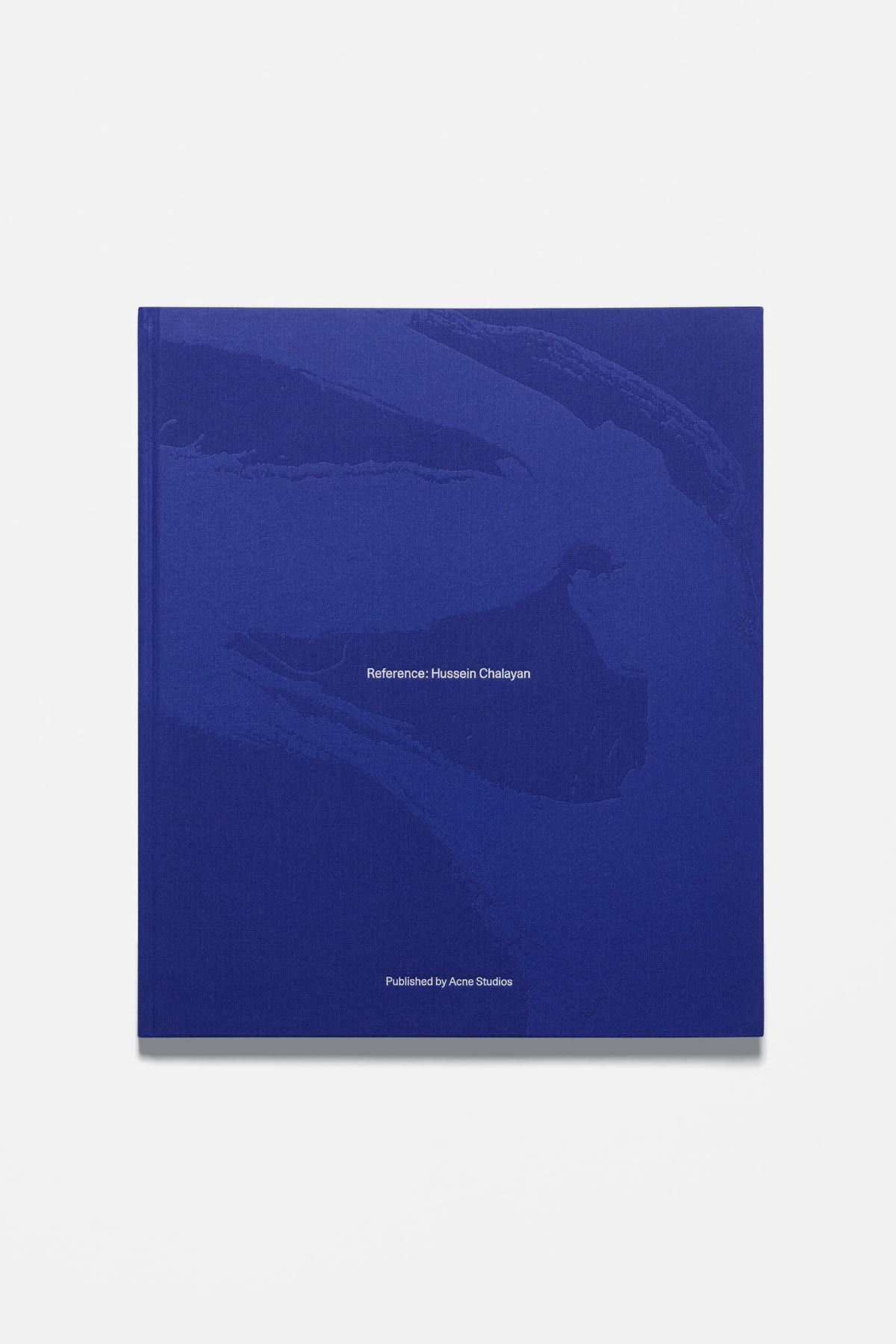 The cover of Reference: Hussein Chalayan, a cobalt blue cloth-bound book with a faint tonal image of a figure, its title in small white type at the centre and "Published by Acne Studios" at the foot