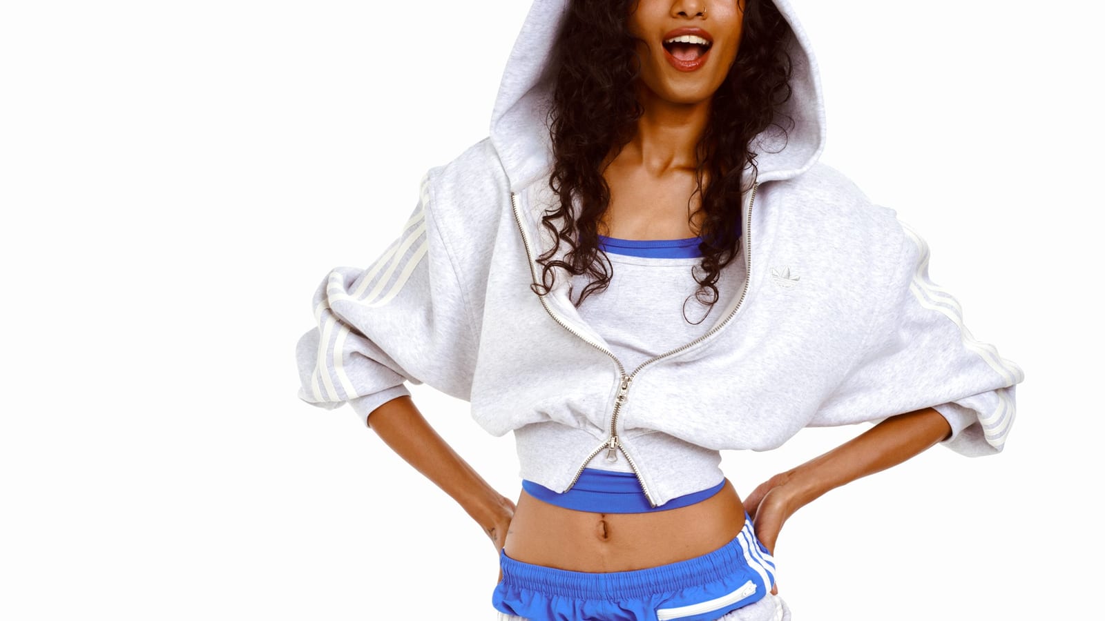 A woman with long curly hair in a cropped grey full-zip hoodie with white stripes on the sleeves, over a grey crop top trimmed in blue and blue track shorts with white stripes