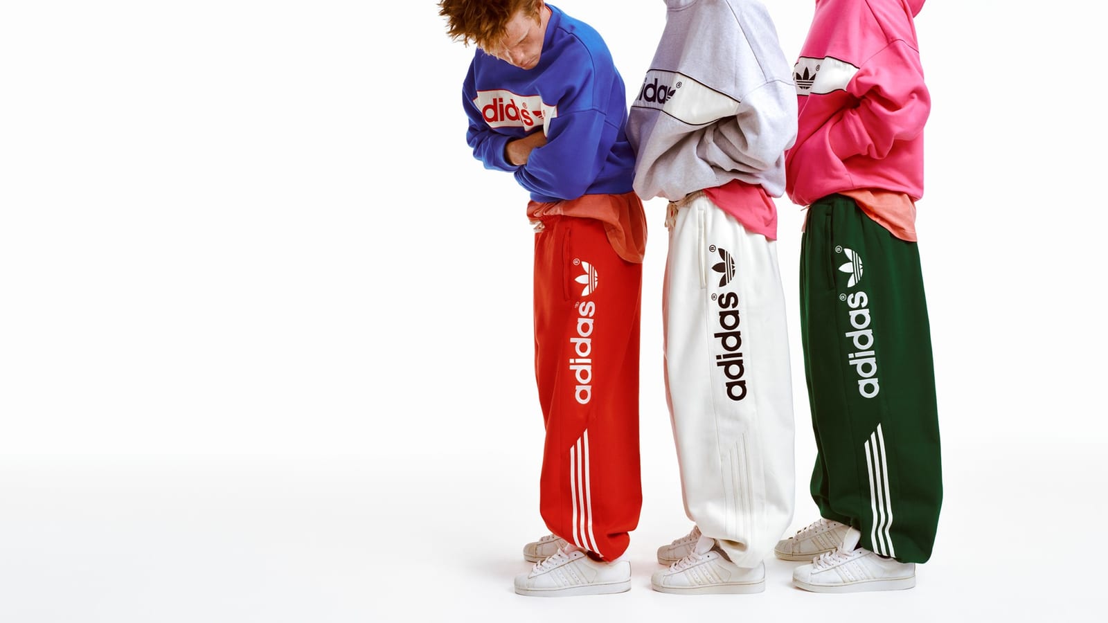 Three models seen from the side in red, white and green sweatpants with vertical adidas Trefoil logos and Three Stripes, under blue, grey and pink sweatshirts and hoodies