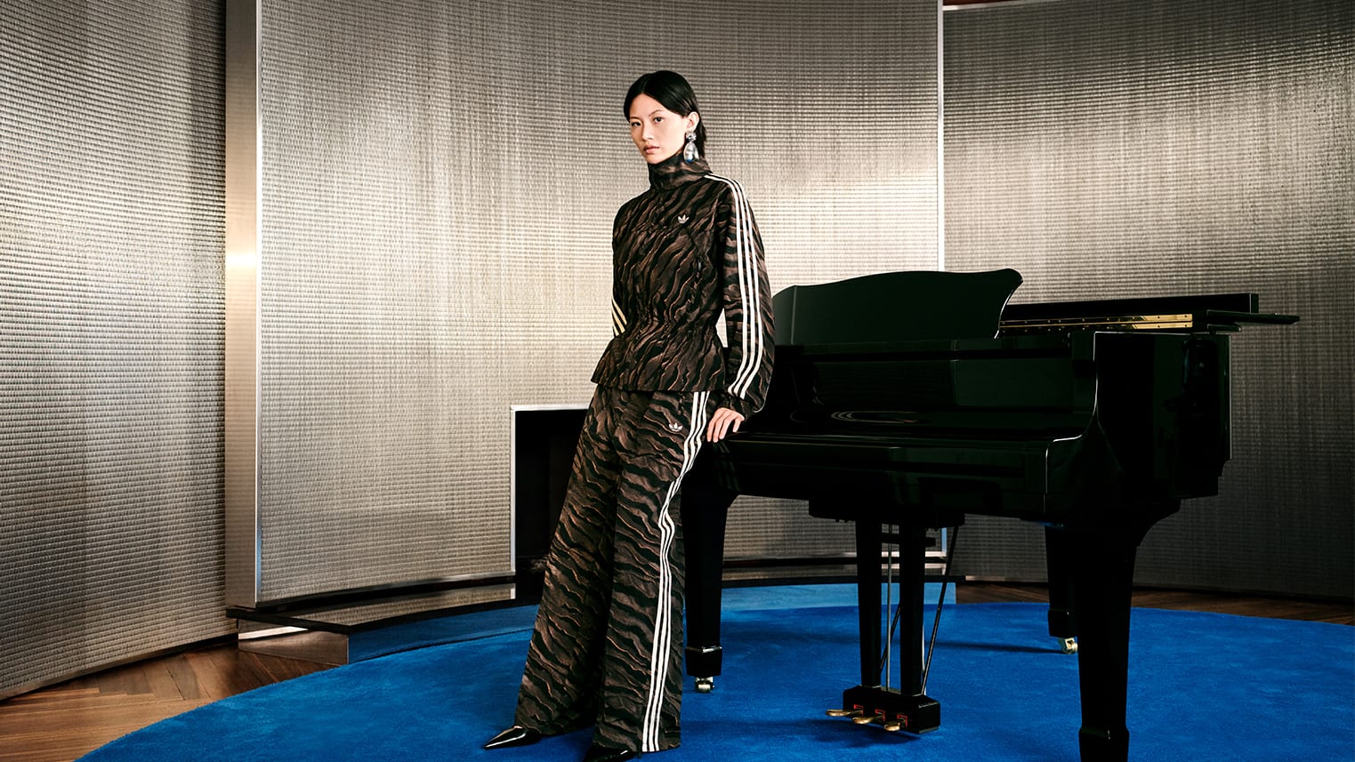 A model leaning on a black grand piano on a round blue rug, in a brown and black wave-print adidas track jacket with a fitted, peplum waist and matching wide track pants, both with cream three stripes