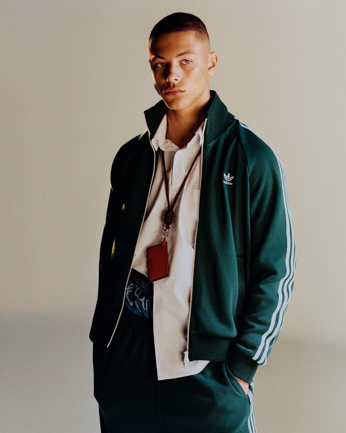 A young man with a short haircut in a dark green adidas track jacket with white three stripes on the sleeves and matching track pants, a white shirt open underneath, and a red leather card holder on a cord around his neck