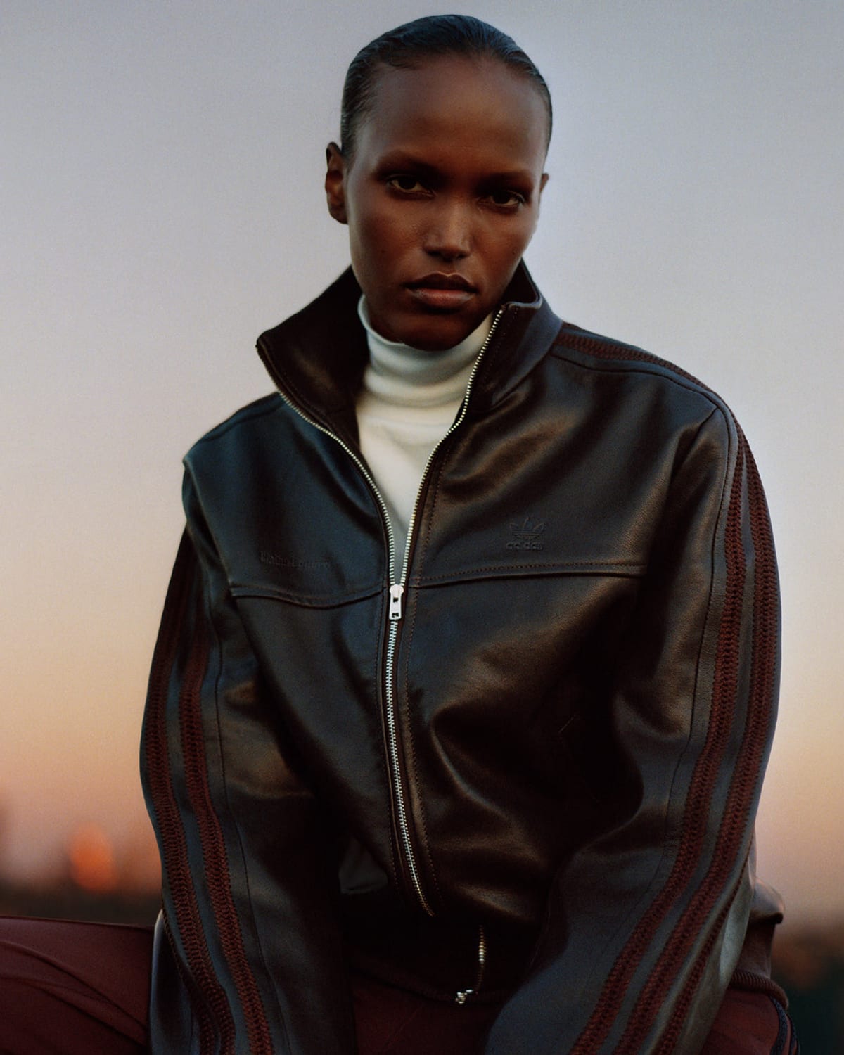A woman with slicked-back hair against a dusk sky in a dark brown leather zip-up track jacket with a stand collar, the adidas trefoil and Wales Bonner name on the chest and textured burgundy stripes down the sleeves, over a white roll-neck