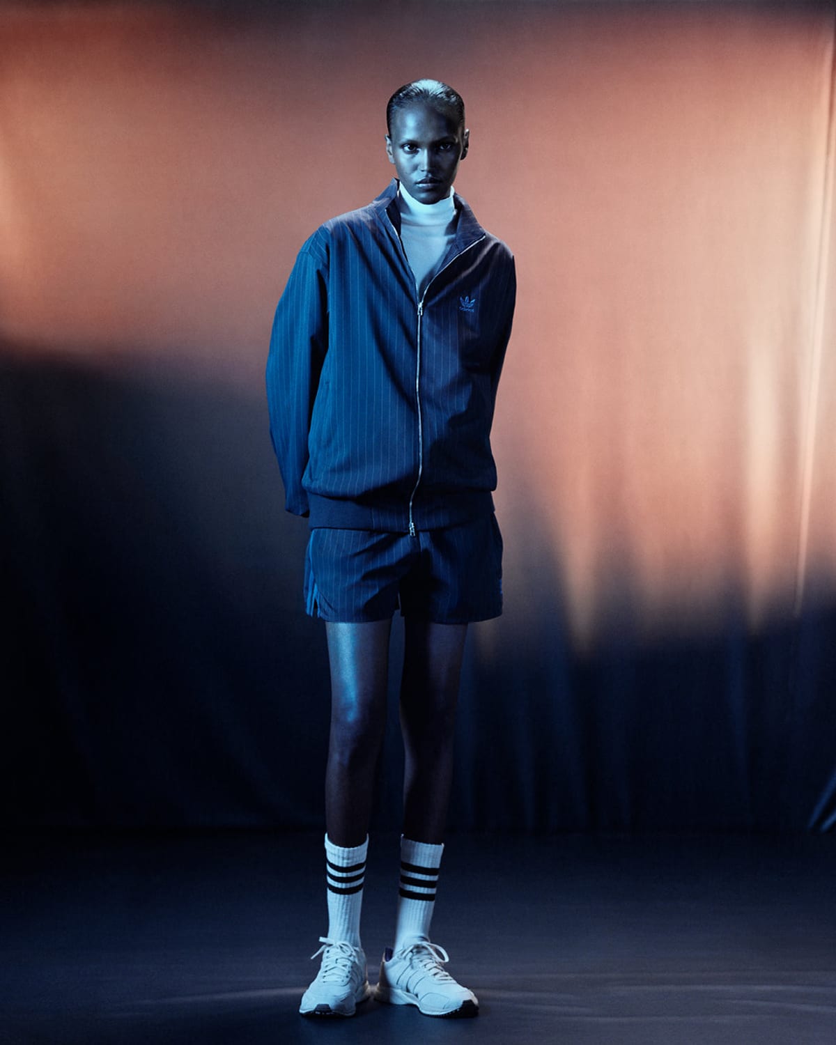 A woman standing in front of a peach and blue backdrop in a navy pinstripe zip track top and matching shorts, a white roll-neck, white socks with black stripes and white trainers