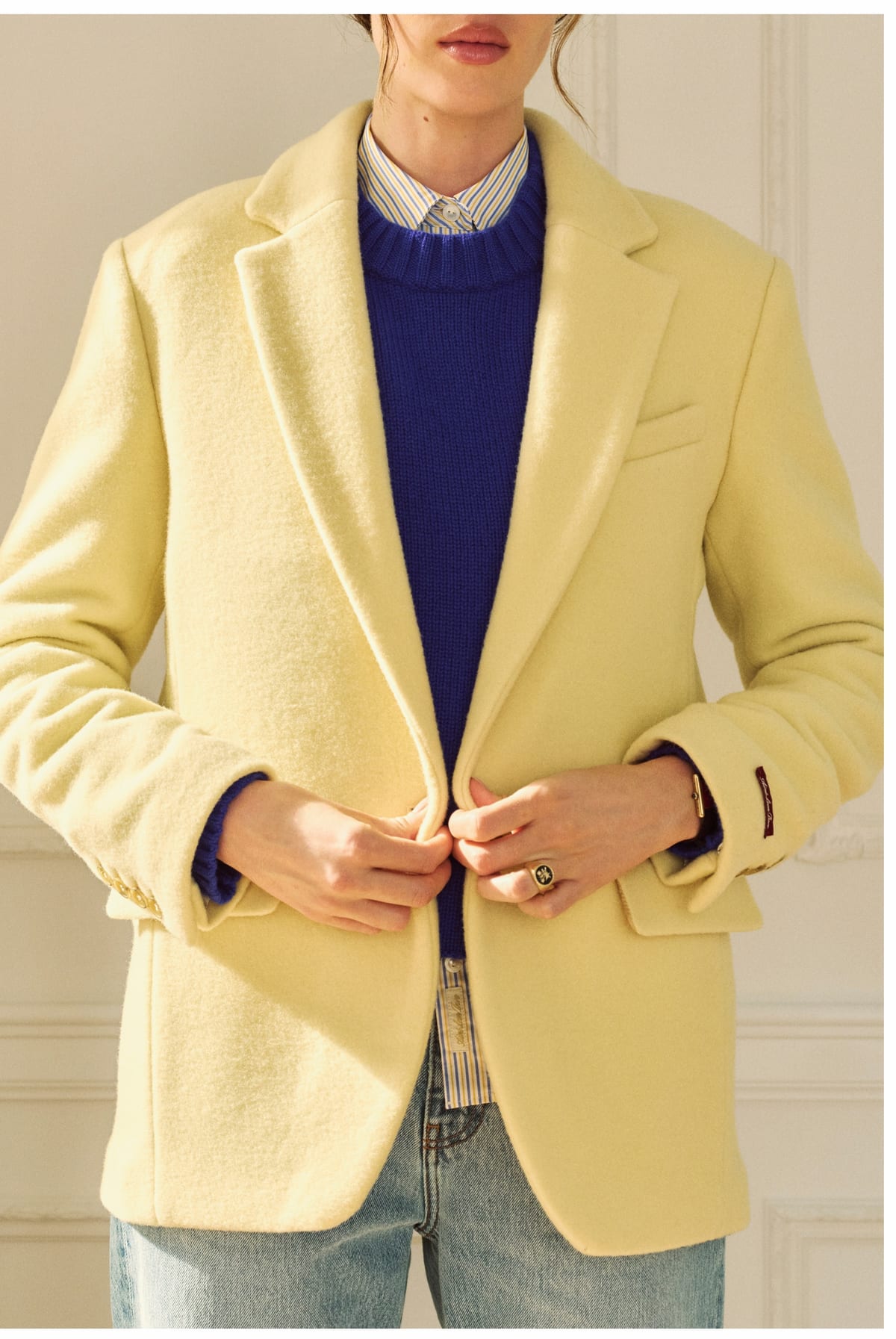 A woman buttoning a pale butter-yellow wool blazer over a cobalt blue crewneck sweater and a striped collared shirt, with light blue jeans, against a cream panelled wall