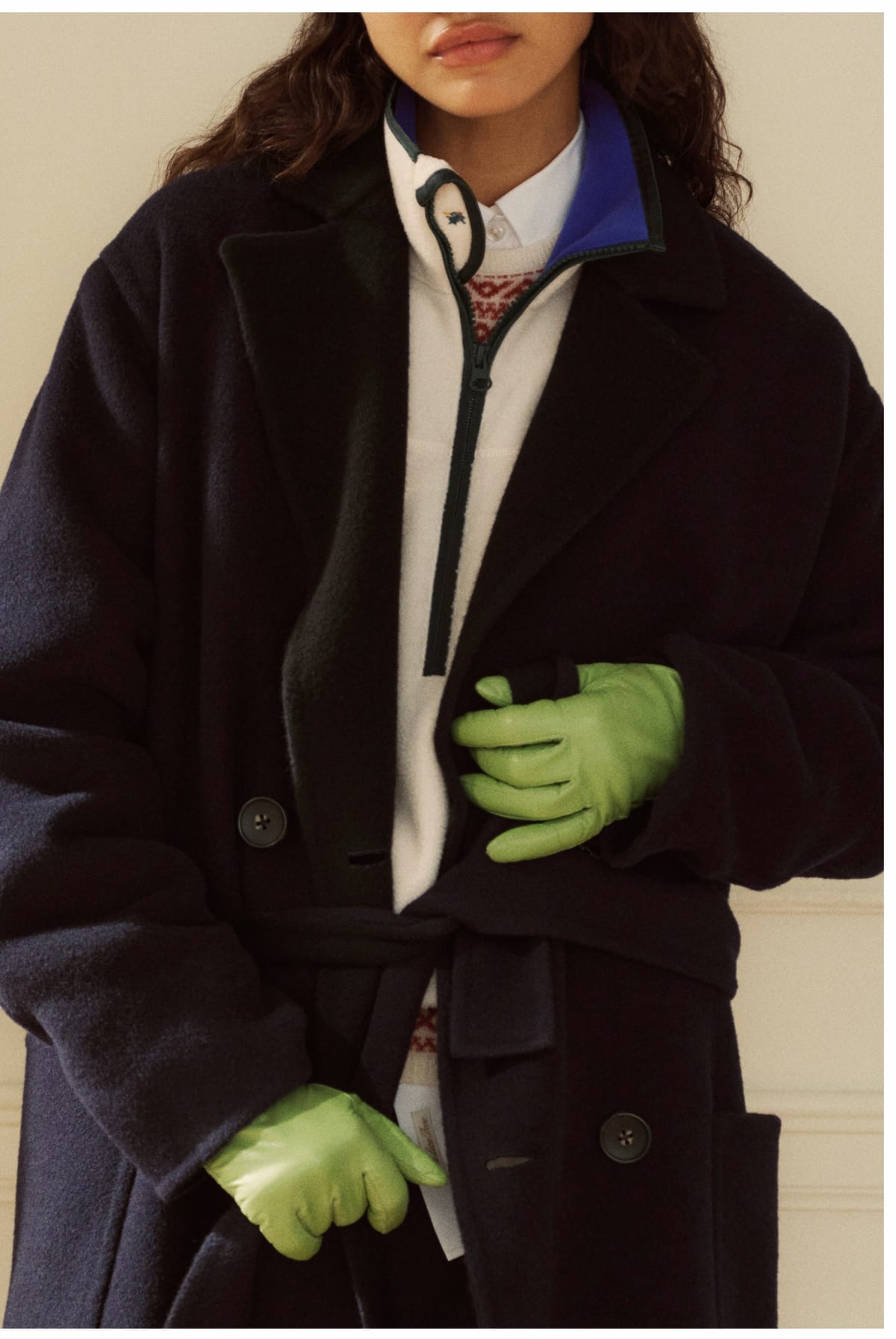 A woman in a long navy double-breasted wool coat, belted loosely at the waist, over a cream half-zip with a blue collar and a patterned knit, wearing bright green leather gloves