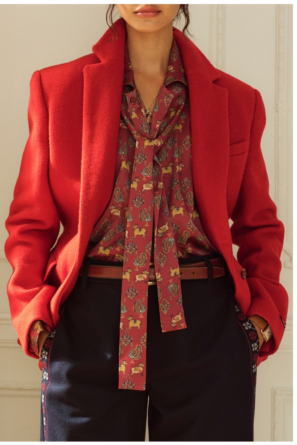 A woman in a bright red wool blazer worn open over a red silk blouse printed with small gold animals and paisleys, its long tie hanging loose, a brown leather belt and dark trousers with an embroidered side stripe