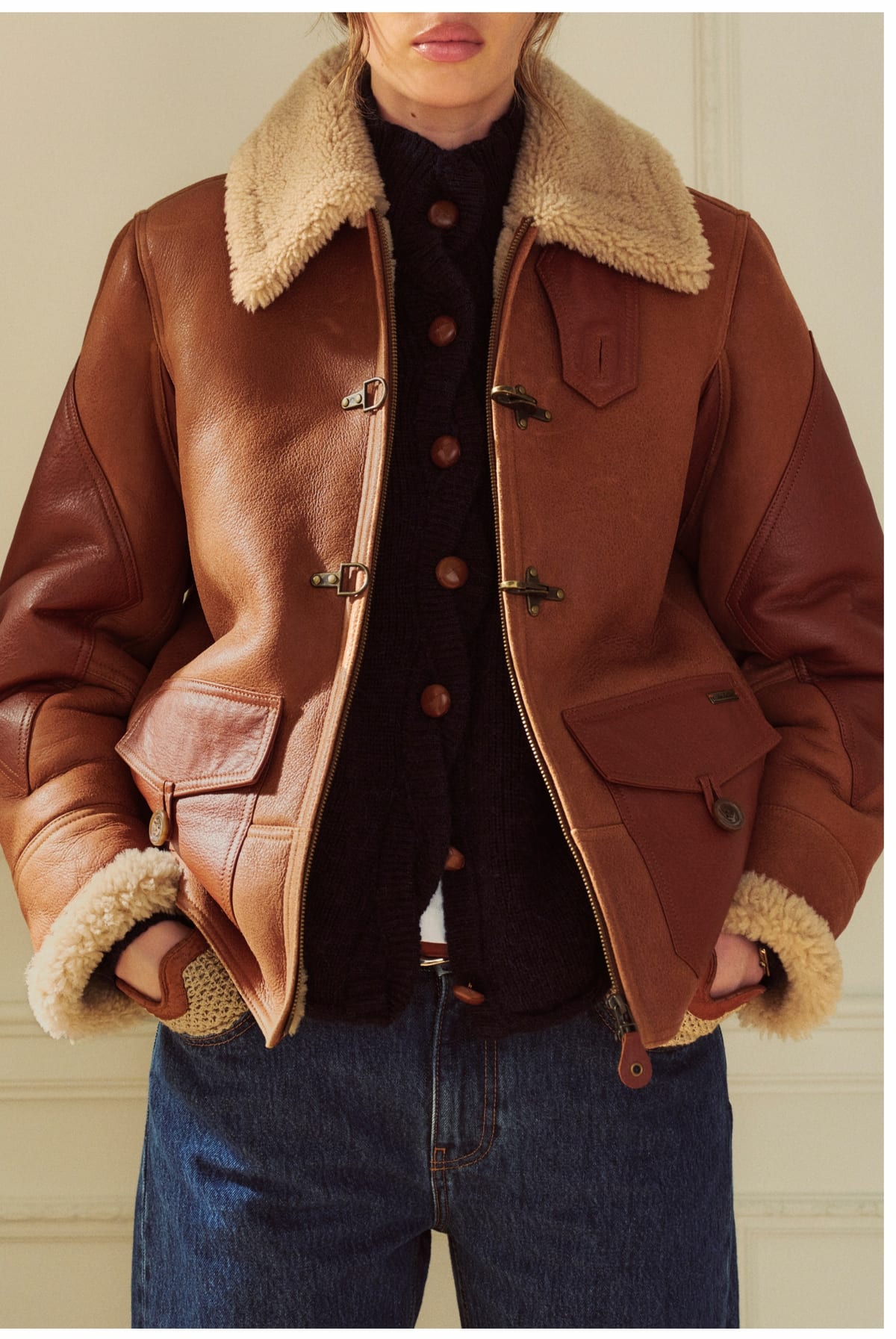 A woman in a brown leather shearling jacket with a cream shearling collar and cuffs, metal buckle clasps and flap pockets, worn open over a dark cable-knit cardigan with brown buttons and dark blue jeans, against a cream panelled wall