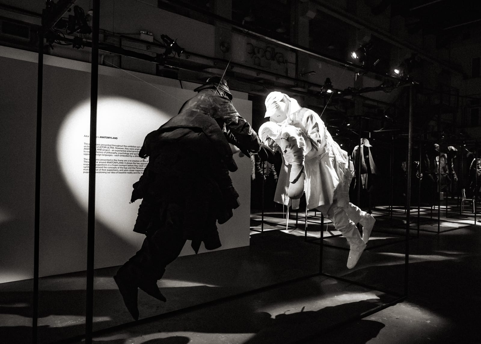 A black-and-white view of two full figures dressed head to toe, with hats and hoods, suspended in mid-air in a dark gallery, one dark and one pale, beside a lit wall of text