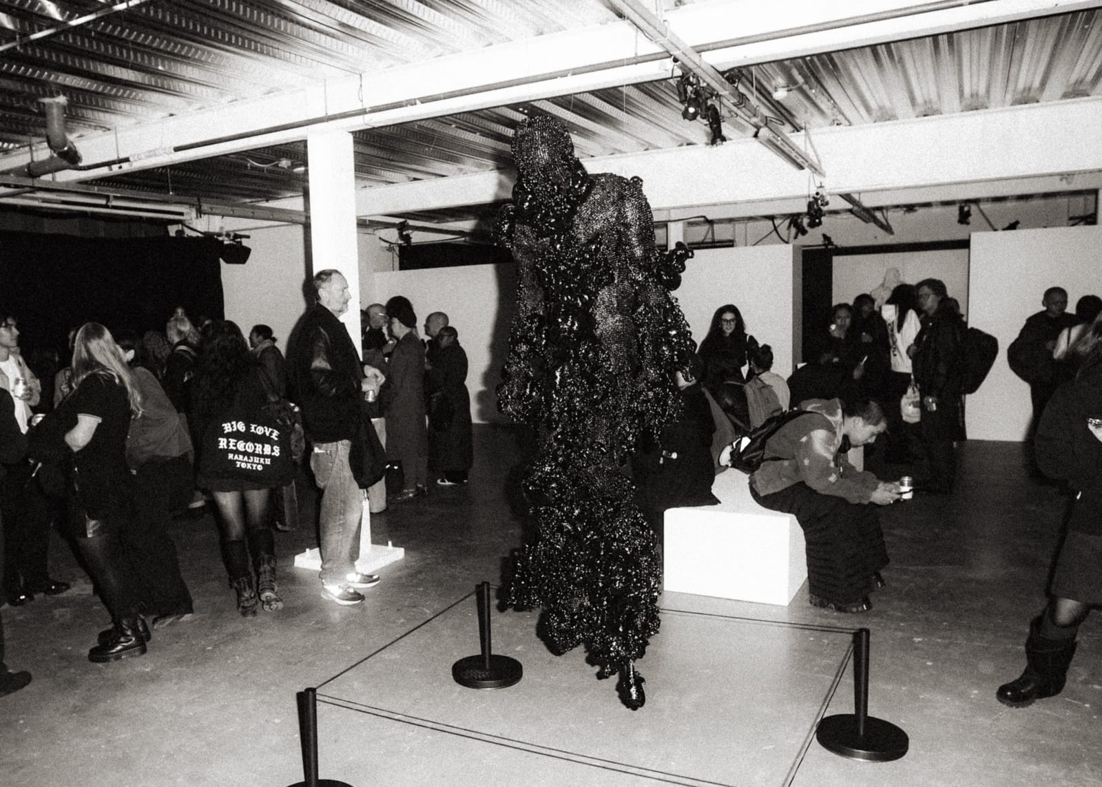A black-and-white view of the exhibition's opening night, with visitors walking among the works and a tall figure in a dark, glittering hooded garment displayed inside a roped-off area, beside a white block where a visitor sits