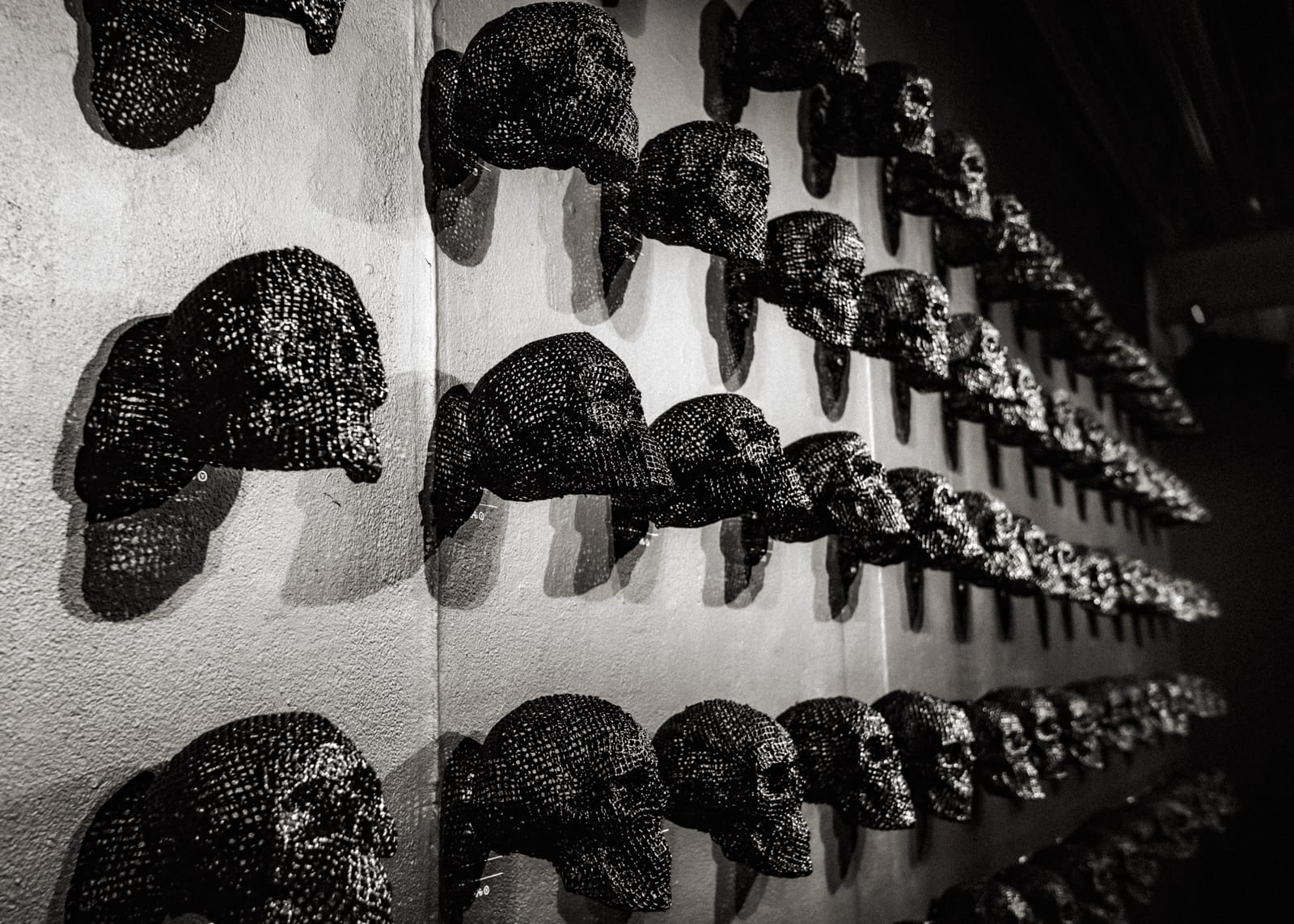 A black-and-white close view of a wall covered in rows of glittering black skull sculptures, receding into the dark