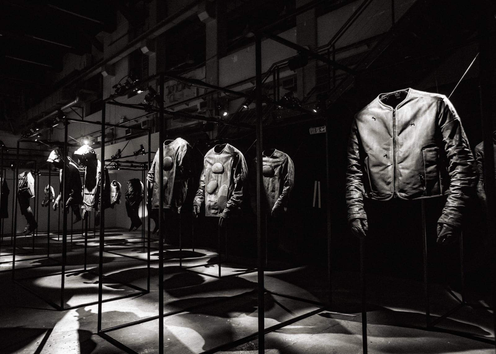 A black-and-white view of a dark exhibition hall where padded leather jackets are mounted inside open metal frames, each lit from above, with a collarless leather zip jacket in the foreground