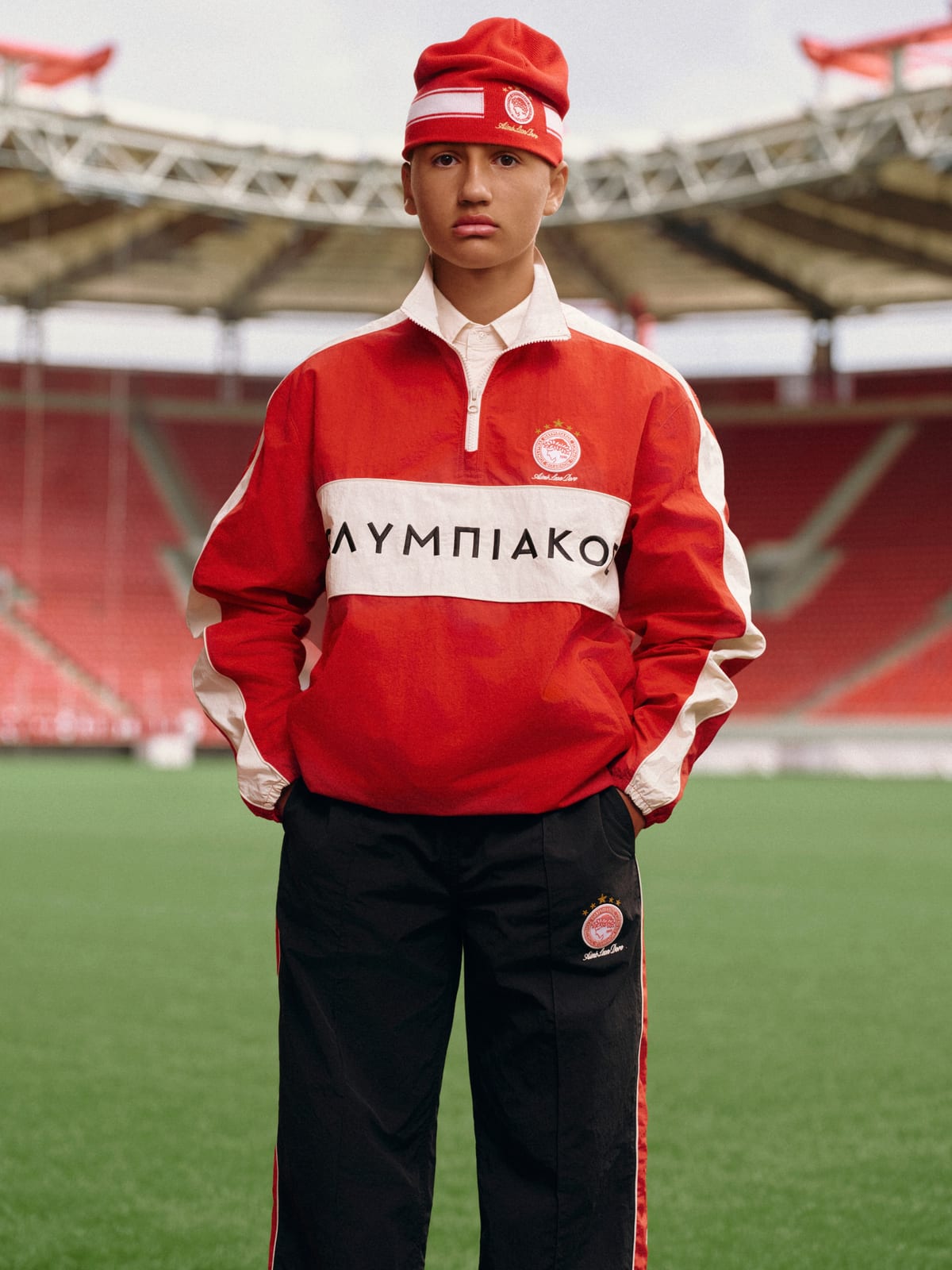 A young man standing on the pitch of an empty stadium with red seats, in a red and cream nylon quarter-zip with ΟΛΥΜΠΙΑΚΟΣ across the chest, a red striped beanie and black nylon track pants with the Olympiacos crest and Aimé Leon Dore script