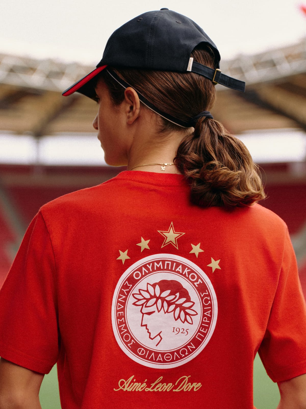 The back of a person with a ponytail and a dark cap, in a red tee printed with a large white Olympiacos crest, five stars and Aimé Leon Dore in script, in front of red stadium seats