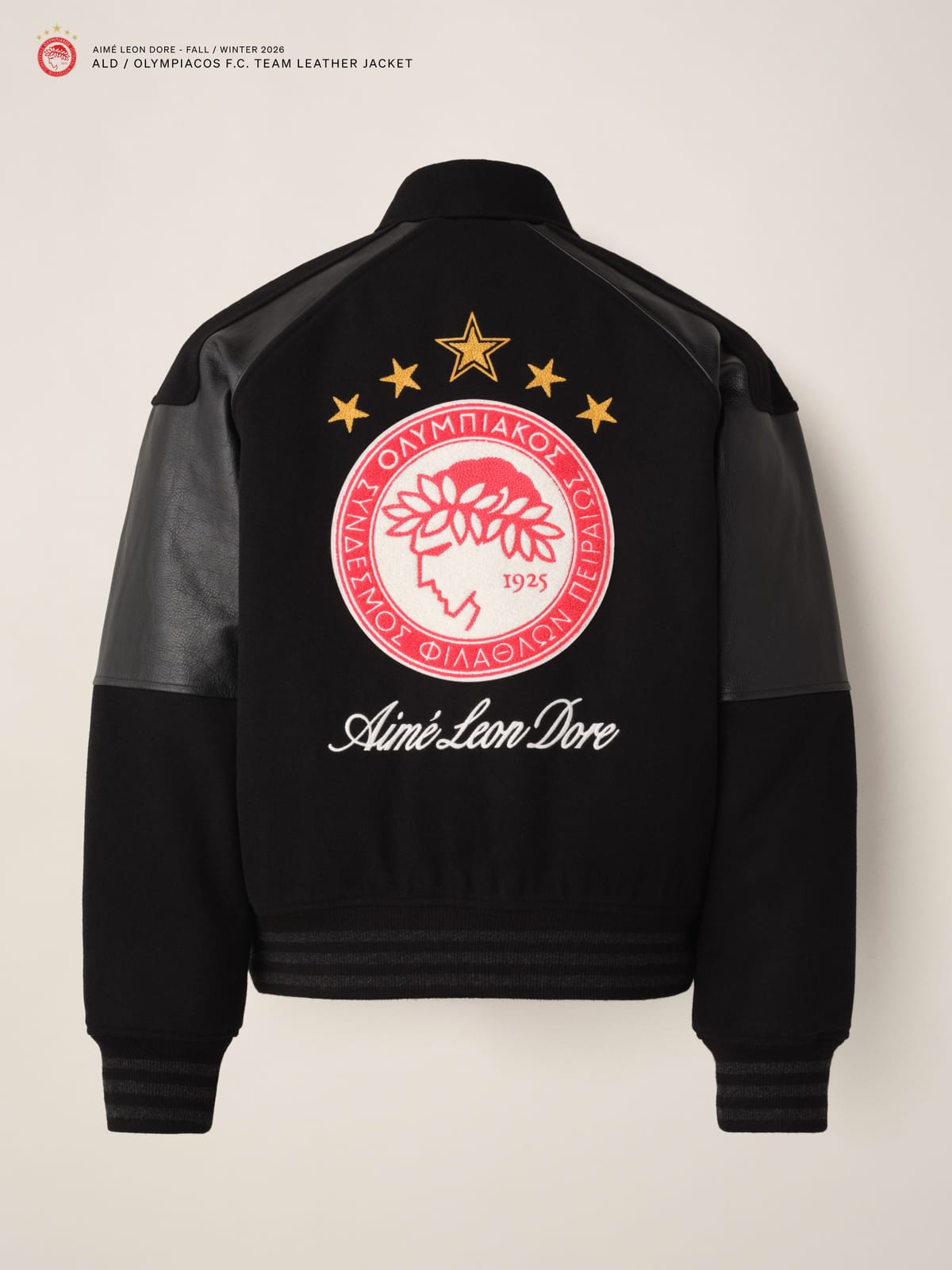 The back of a black wool varsity-style jacket with black leather over the shoulders and upper sleeves, a large red and white Olympiacos crest under five gold stars, and Aimé Leon Dore in white script below it, on a cream background