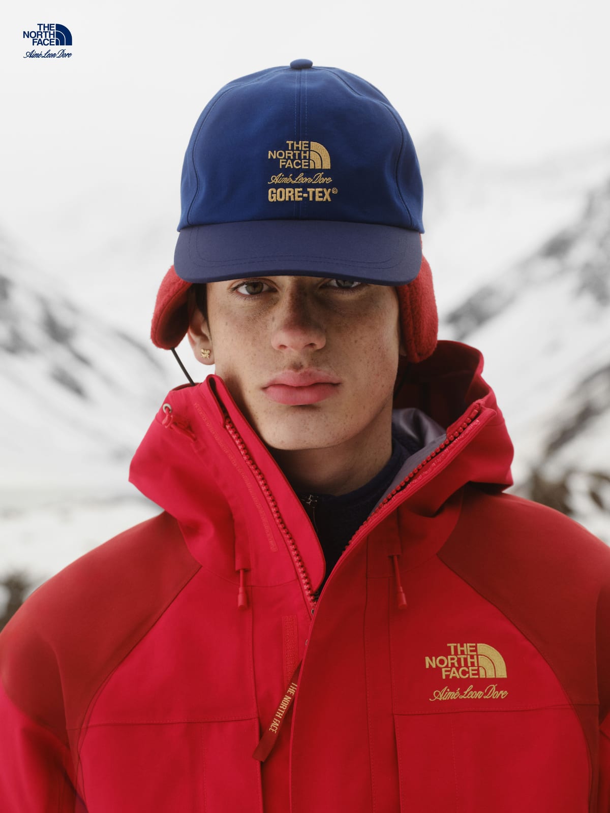 A model in the red GORE-TEX 2000 Mountain Jacket, open at the collar over a navy layer, with gold The North Face and Aimé Leon Dore embroidery at the chest, under a navy GORE-TEX cap with red ear flaps showing beneath it