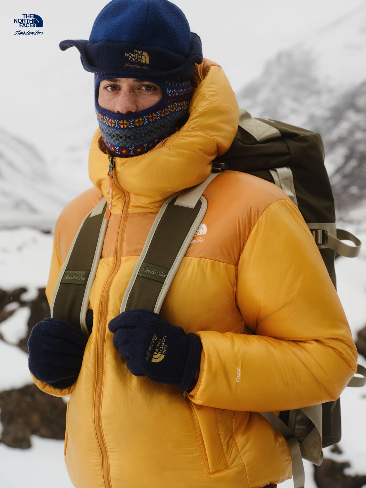 A model in the gold Reversible Pertex Nuptse Jacket with a Fair Isle balaclava, a navy fleece earflap hat and navy fleece gloves, carrying a large olive pack in front of snowy mountains
