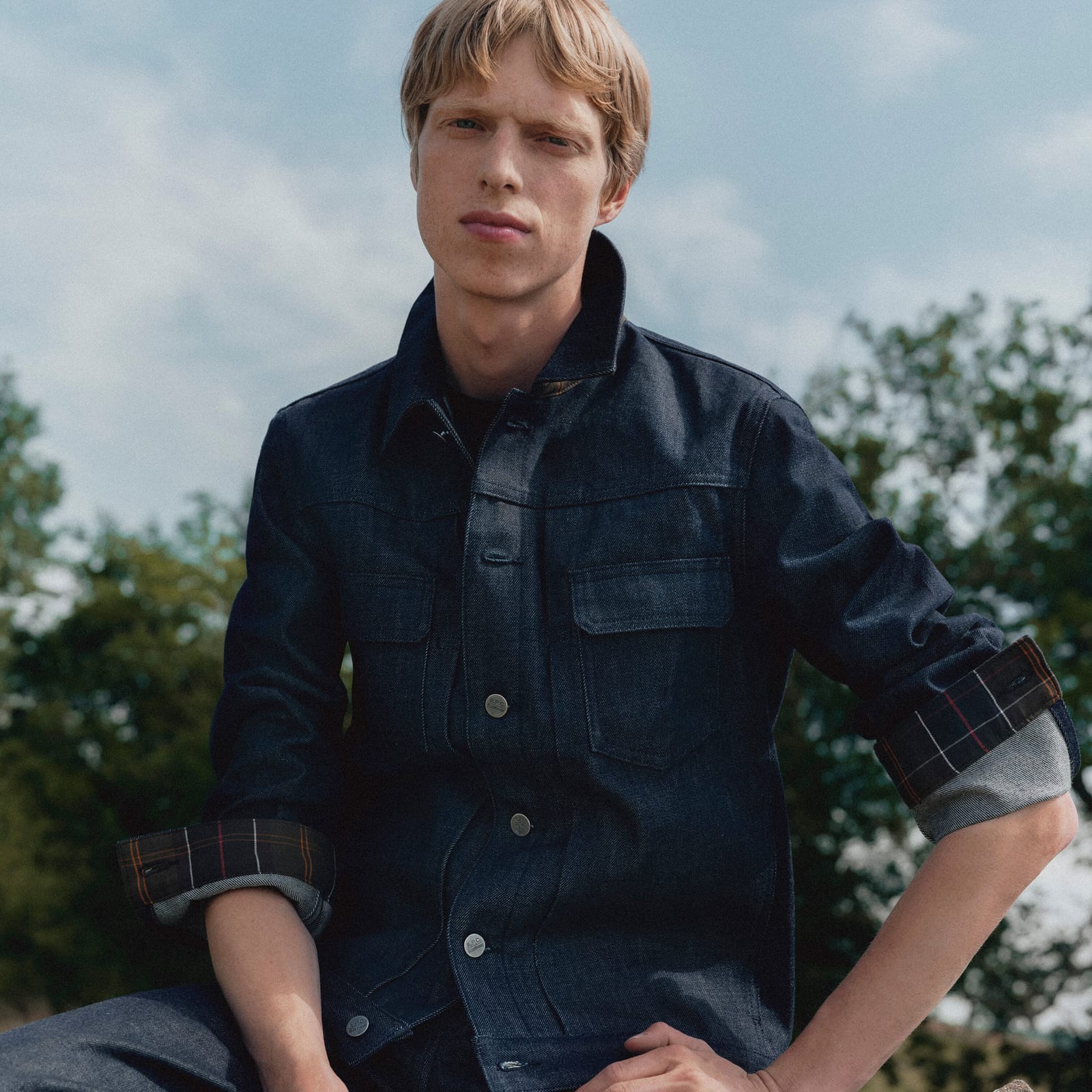 A young man with blond hair in an indigo denim trucker jacket with two chest pockets and metal buttons, sleeves rolled to show a tartan lining, sitting outdoors against trees and a cloudy sky