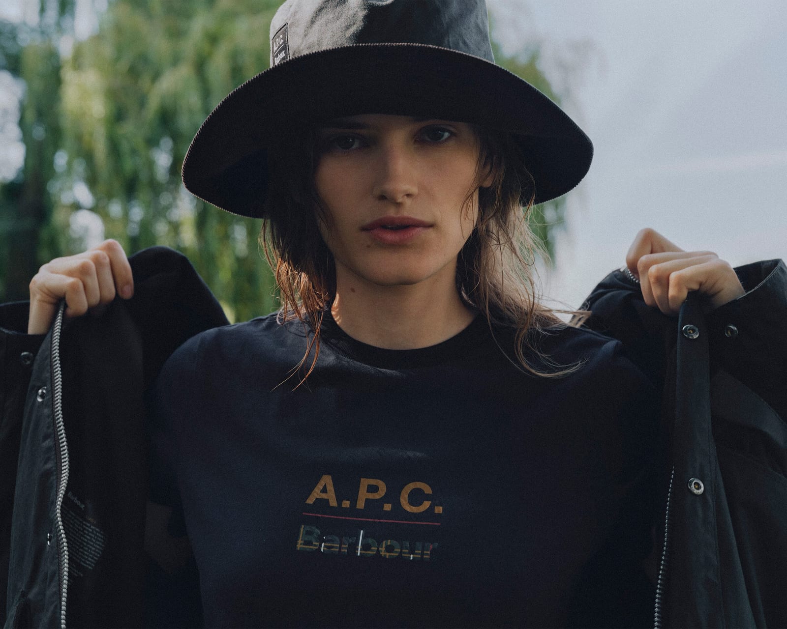 A model in a black waxed bucket hat holding open a dark jacket over a black T-shirt printed with A.P.C. in gold and Barbour beneath it in tartan-filled letters