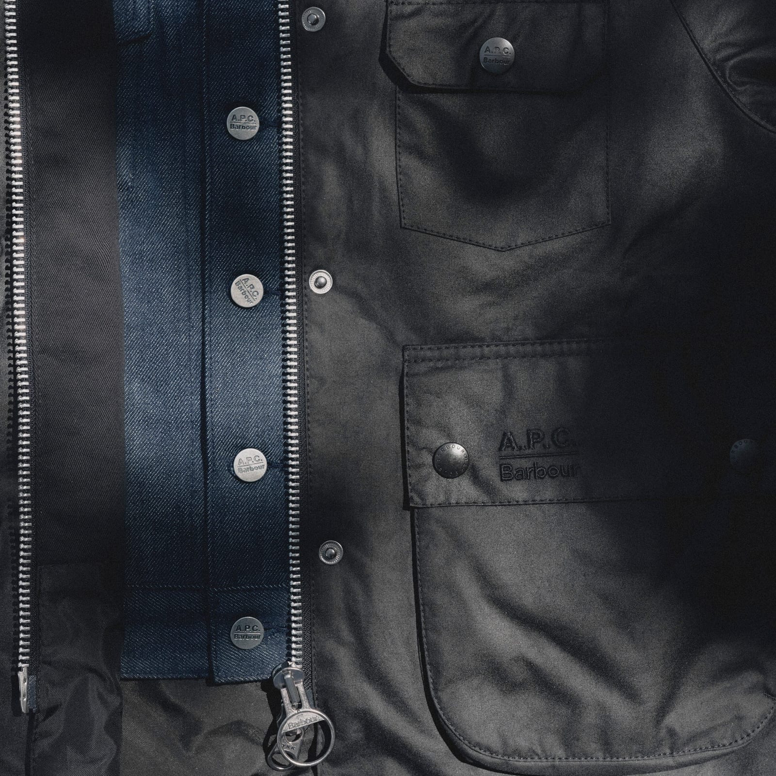 Close-up of a dark waxed jacket worn open over an indigo denim jacket: both have metal buttons stamped A.P.C. Barbour, and the waxed jacket's flap pocket carries a tonal A.P.C. Barbour embroidery