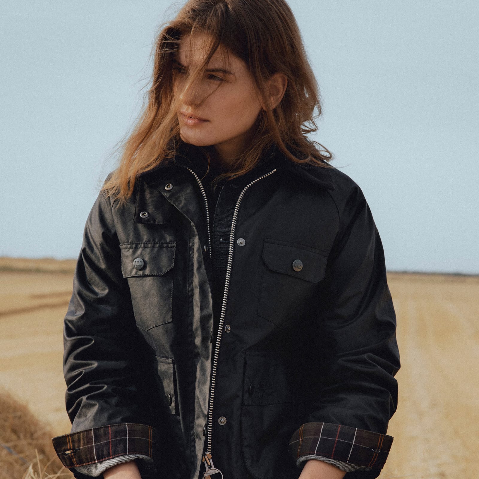 A woman with long windswept hair standing in a harvested wheat field in a black waxed jacket with a silver zip, two snap-flap chest pockets and its sleeves turned back to show a dark tartan lining