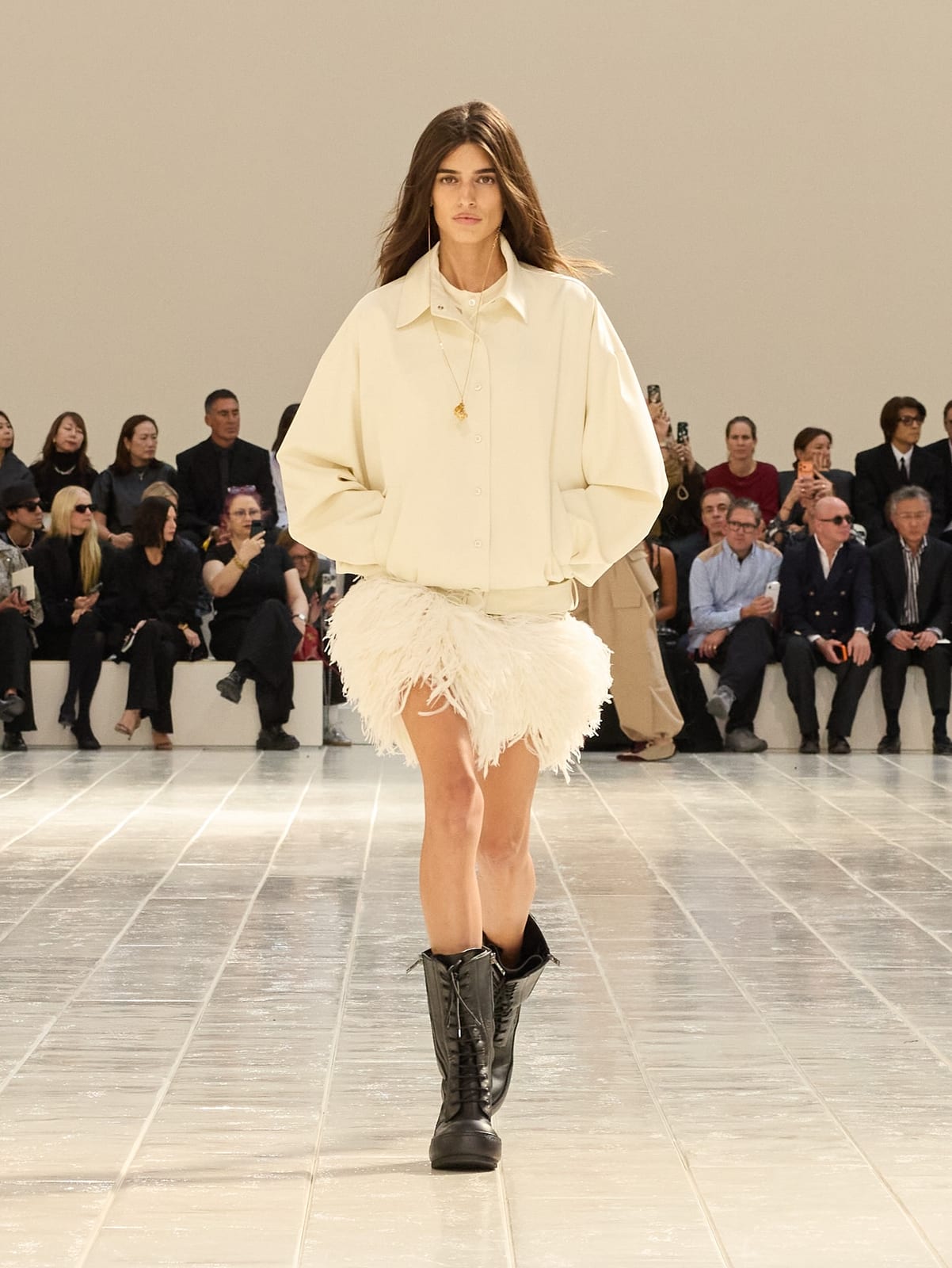 A model on Balenciaga's glossy white runway in a cream bomber jacket, a white feathered mini skirt and black lace-up boots, with the front row behind her