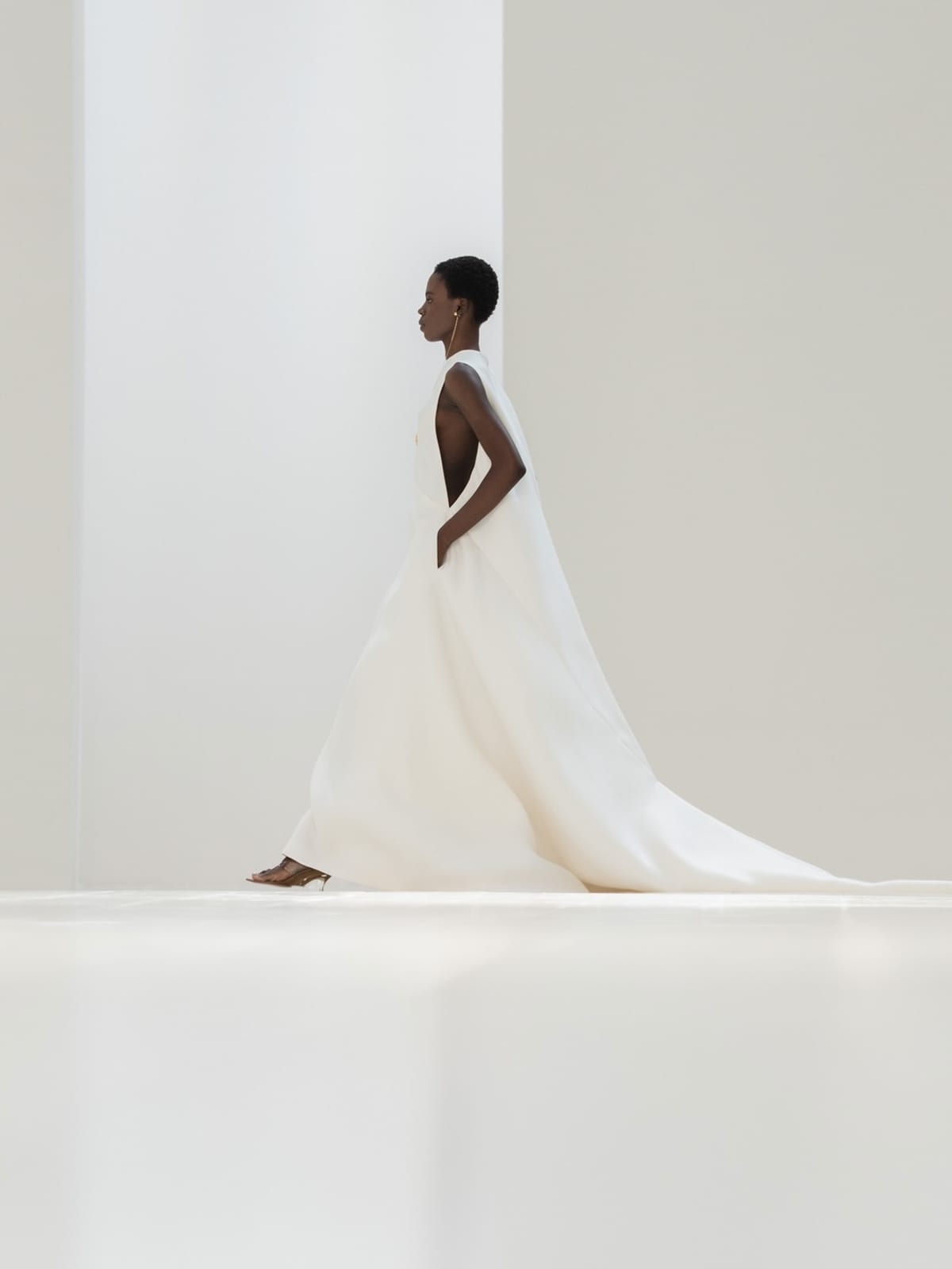 A model in profile on the white Balenciaga runway wearing a sleeveless white gown with a long train