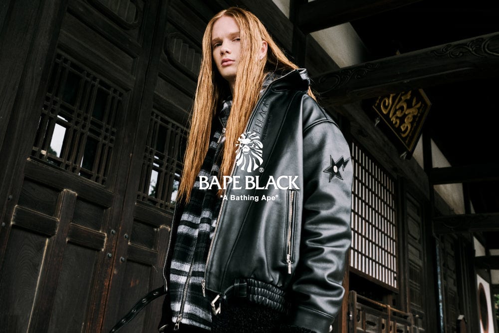 A model with long red hair in a black leather biker jacket over a black-and-white plaid shirt, in front of dark wooden doors, with the BAPE BLACK logo across the chest