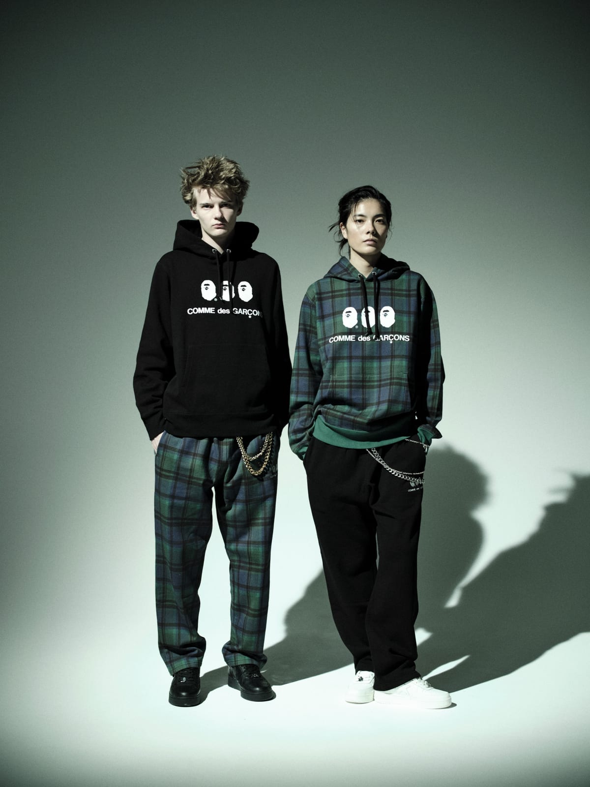 Two models in the BAPE x Comme des Garçons fall/winter 2026 collection: a black hoodie with three ape heads above the COMME des GARÇONS logo, and a green Black Watch check hoodie, both worn with check or black sweatpants and wallet chains