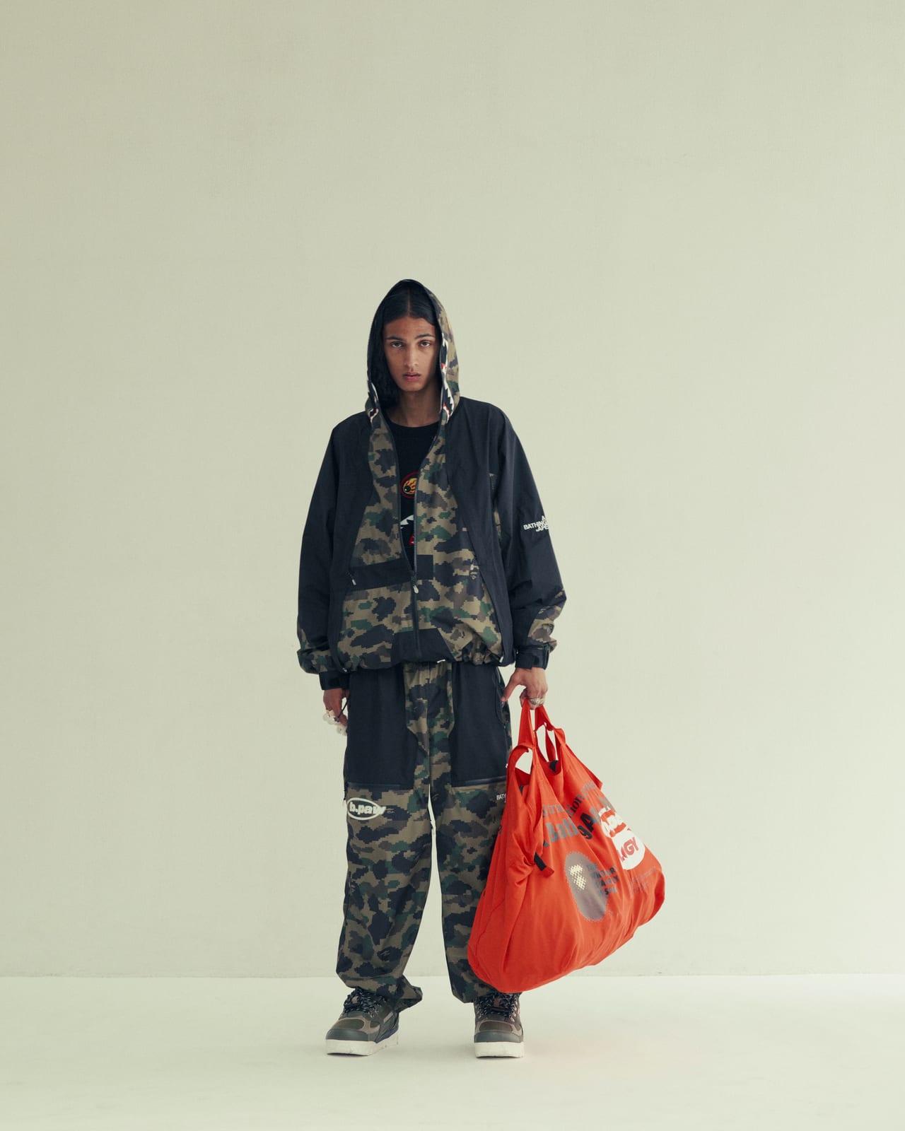 A model in a black hooded jacket and wide trousers in BAPE's green 1st Camo with black panels, carrying a bright orange tote bag, in a pale studio