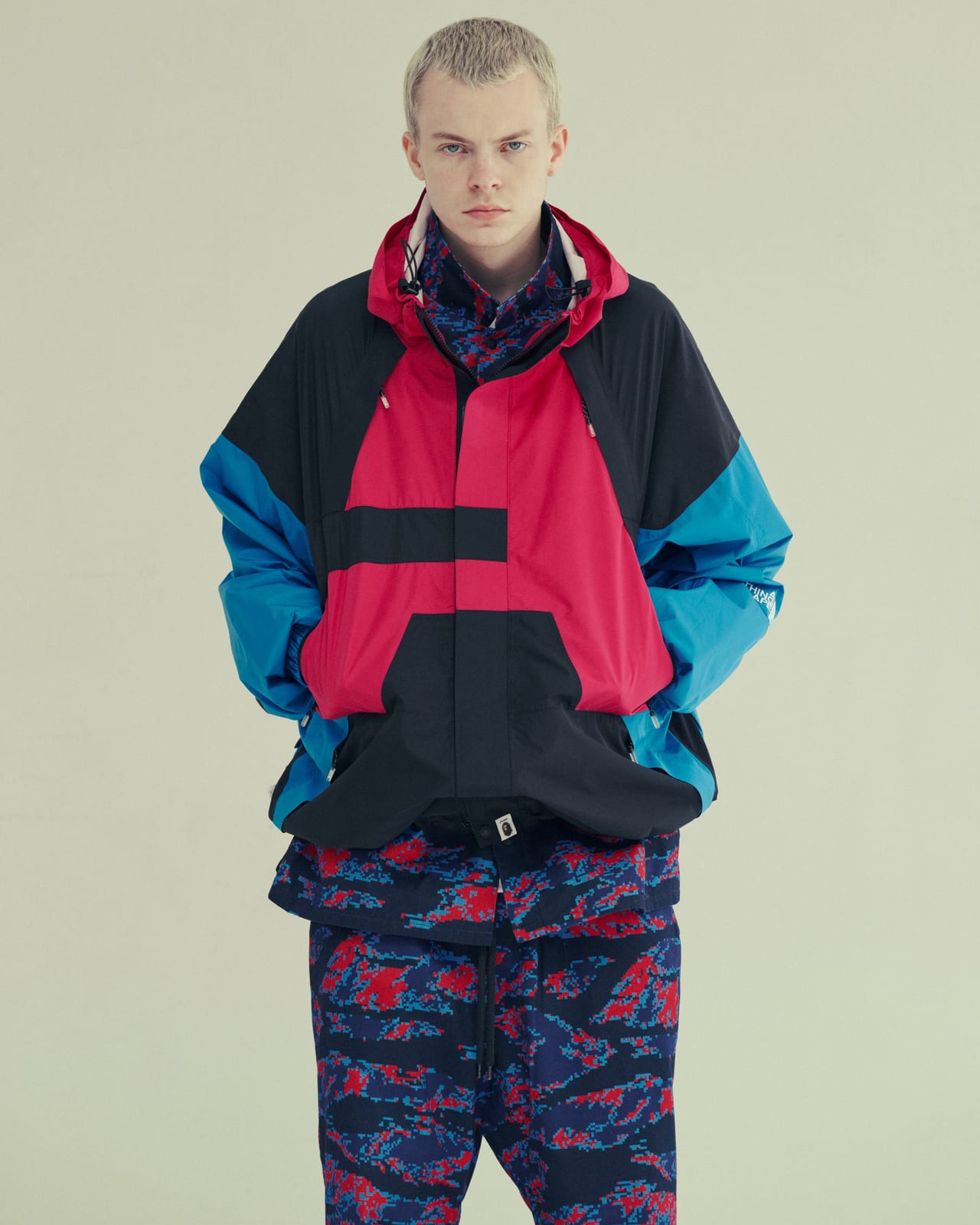 A model with cropped bleached hair in a BAPE hooded anorak in black, magenta and bright blue colour blocks with a drawcord hem and chest pockets, over blue and red tiger-camo trousers, against a pale green studio wall