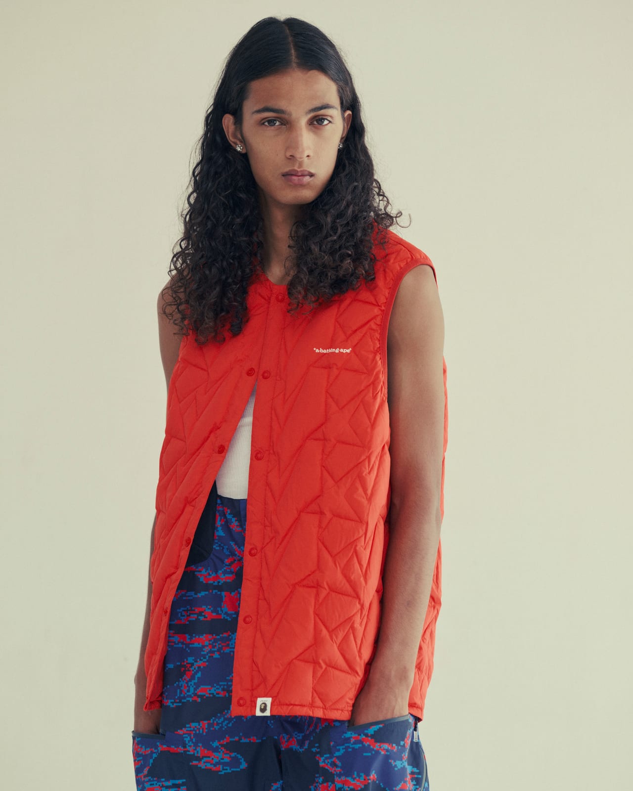 A model with long dark hair in a bright orange quilted vest over a navy jacket, with a small ape-head label at the hem, over navy and blue-and-red digital tiger-camo trousers, standing in a pale studio