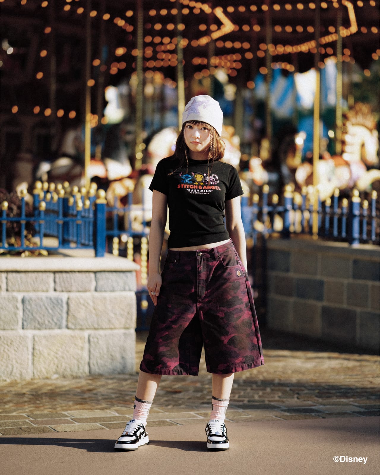 A model in a white star beanie, a black baby tee printed STITCH & ANGEL with Stitch, Baby Milo and Angel, and wide purple camo shorts in front of a carousel