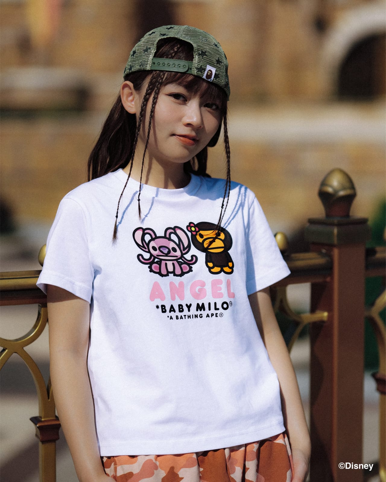 A model in a green star-mesh cap and a white T-shirt printed with Angel, Baby Milo with a flower, and the words ANGEL, BABY MILO and A BATHING APE
