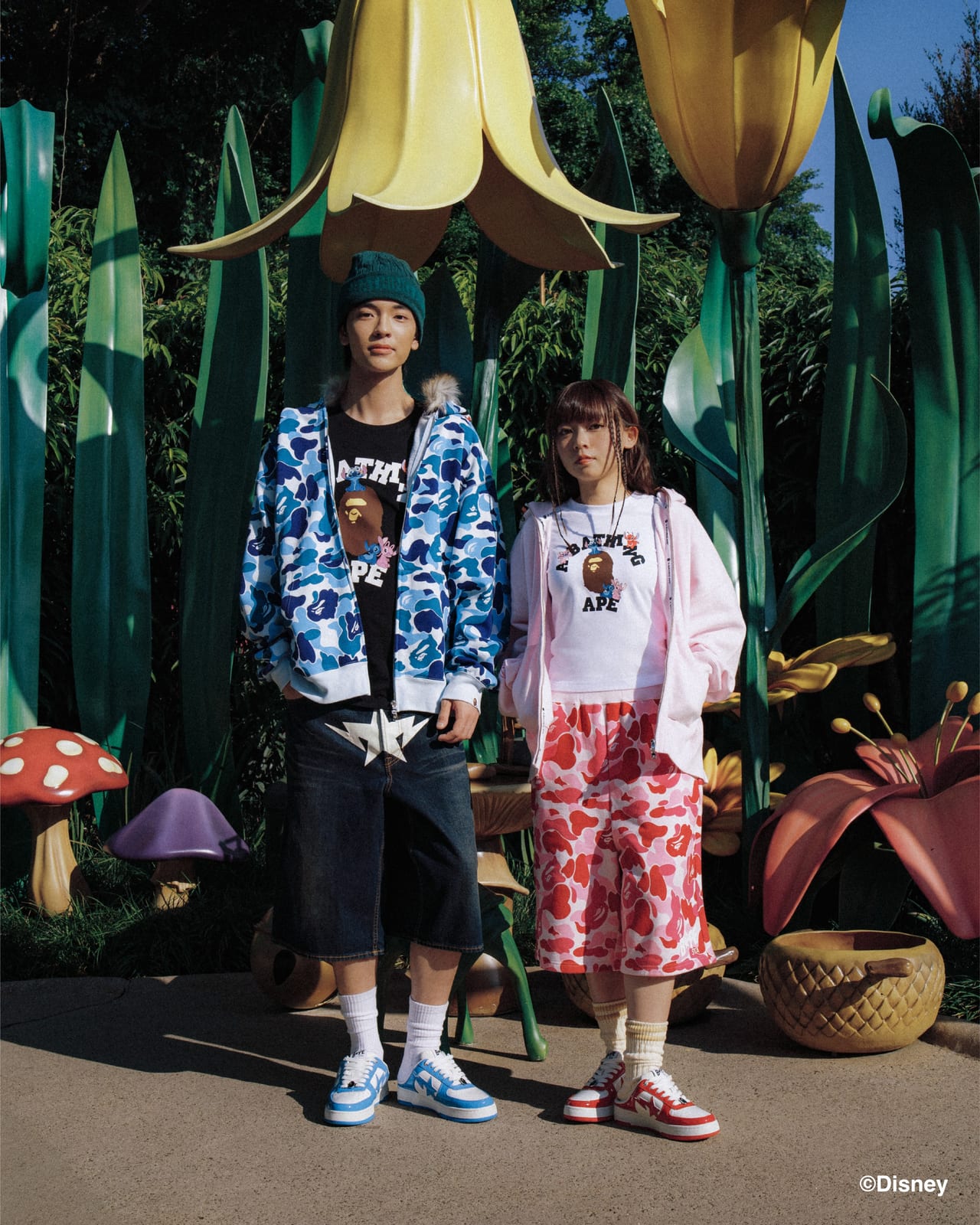 Two models among oversized garden props wearing BAPE x Stitch college-style tees under a blue camo zip hoodie and a pale pink hoodie, with pink and red camo shorts