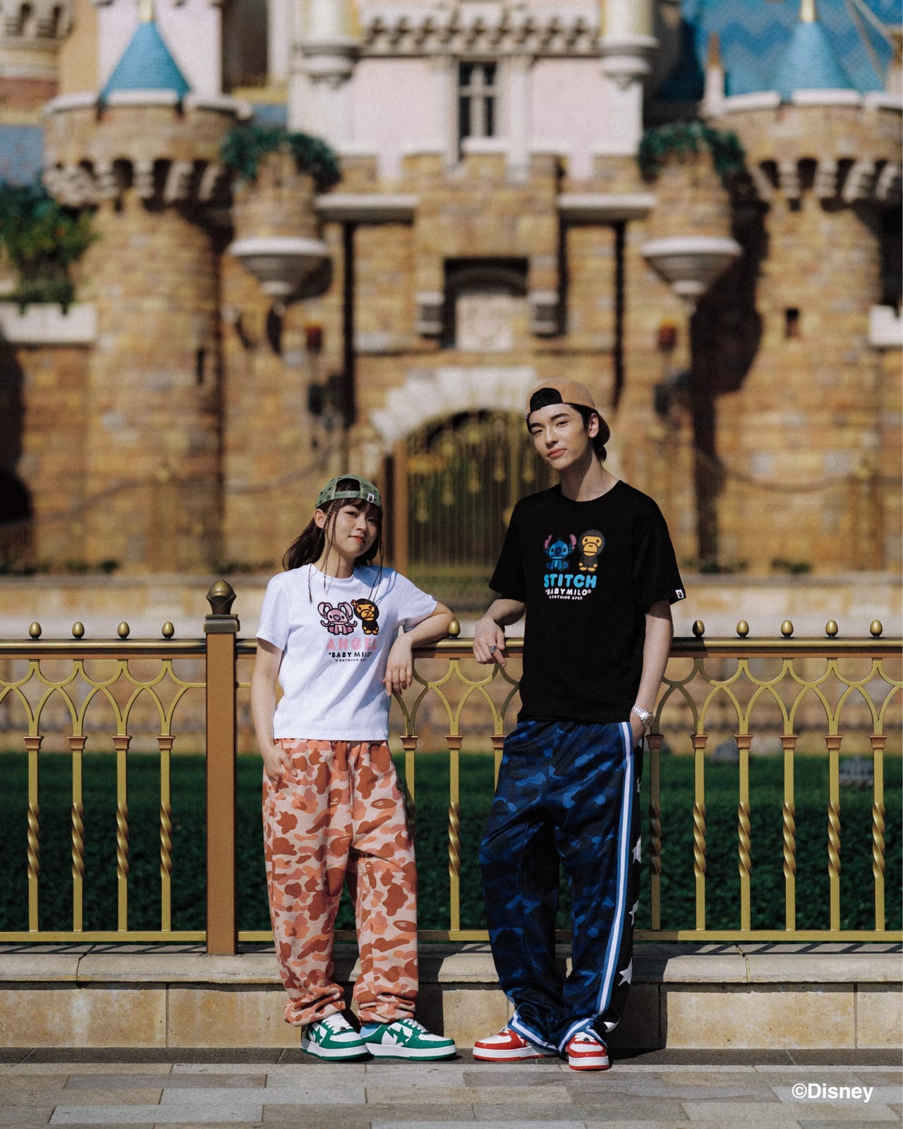 Two models in front of a castle wearing BAPE x Stitch T-shirts: a white tee with Angel and Baby Milo, and a black tee with Stitch and Baby Milo, over camo pants