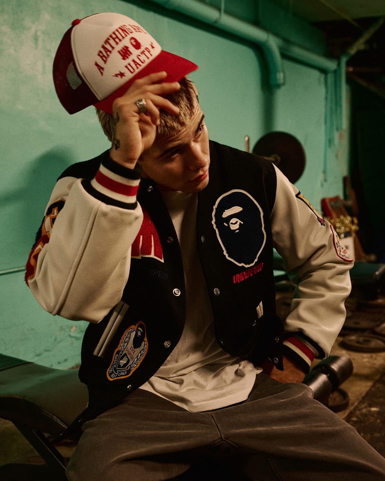 A man in the BAPE x UNDEFEATED varsity jacket and a red and white A Bathing Ape mesh cap, one hand on his head, sitting against a green wall