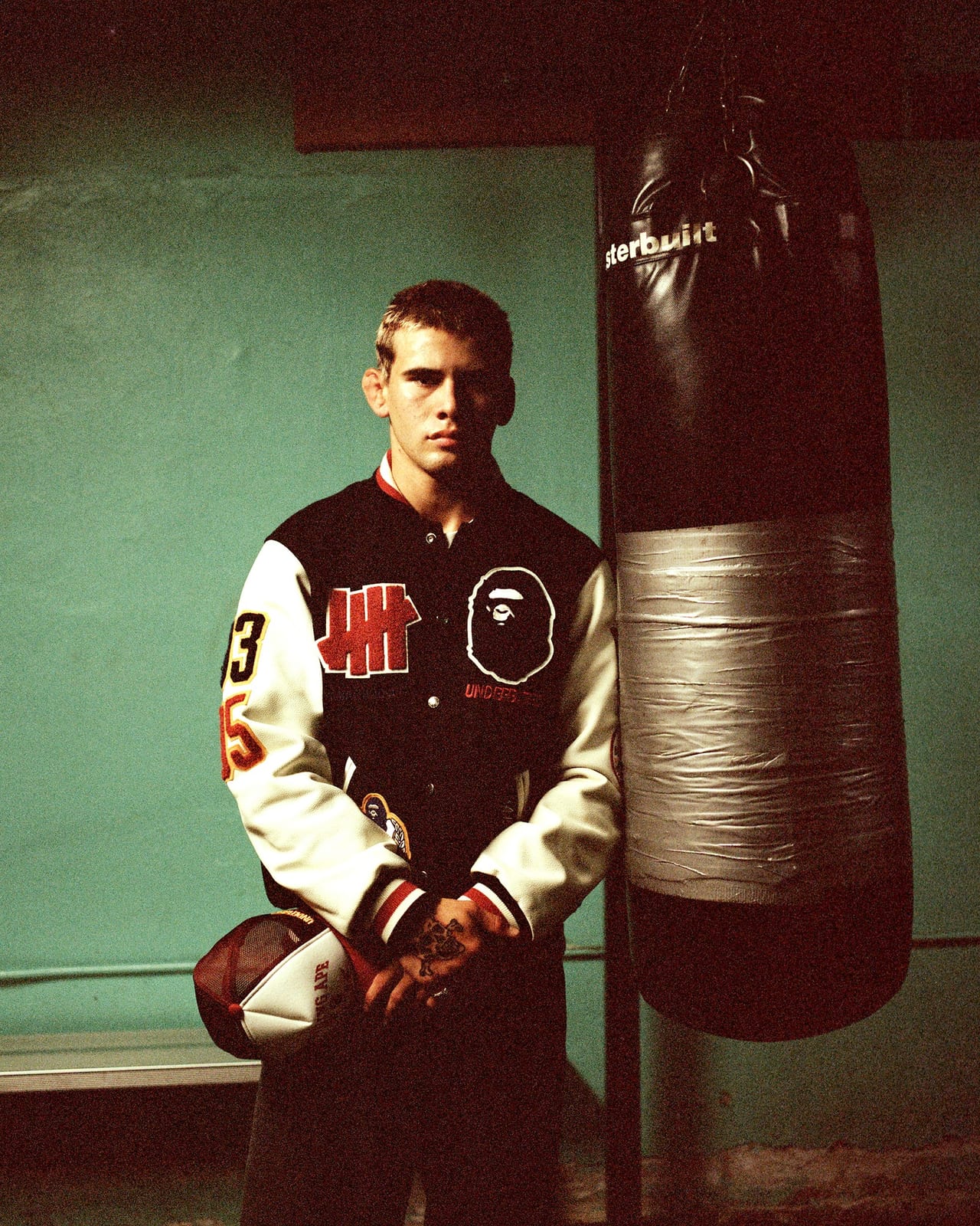 A young man in the black BAPE x UNDEFEATED varsity jacket with cream leather sleeves, the UNDEFEATED 5-Strike in red and the BAPE ape head on the chest and 93 and 05 on the sleeve, holding a mesh cap beside a punching bag in a green-walled gym