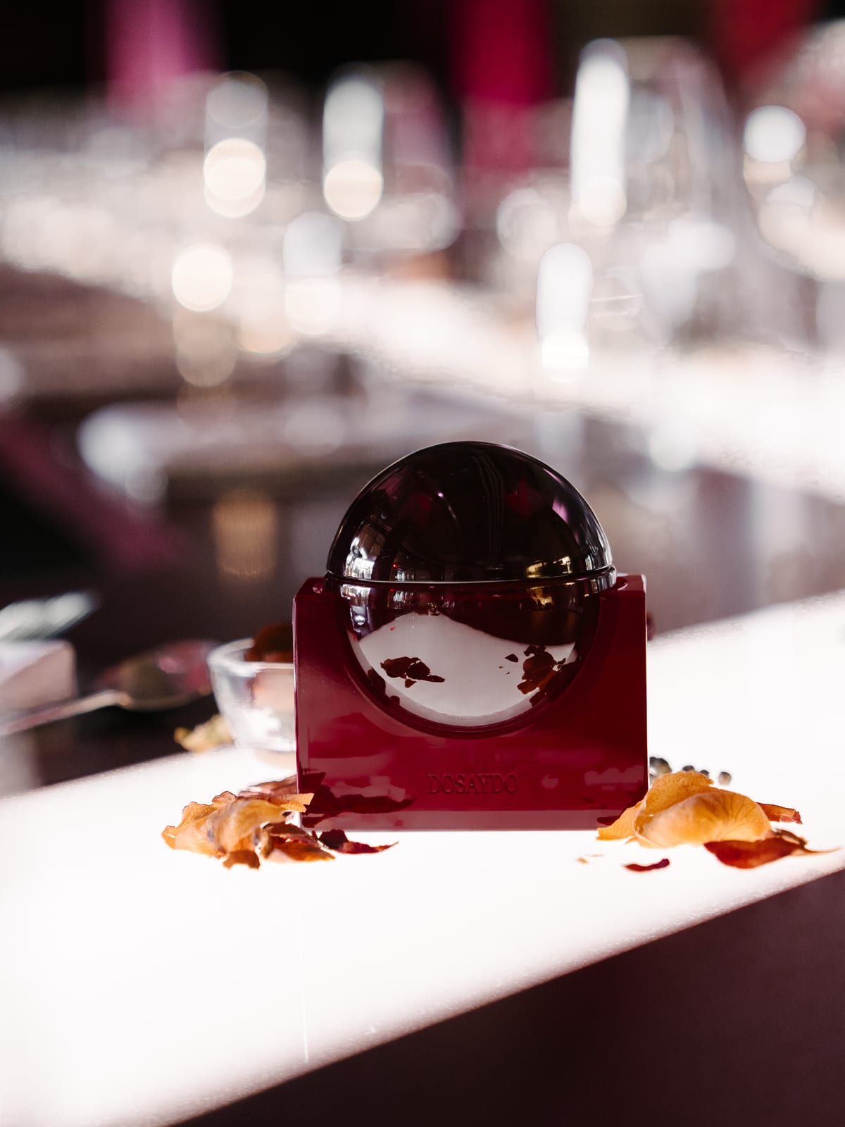 A DOSAYDO bottle in its red block stand on a lit white dinner table, with dried leaves around it and blurred wine glasses behind