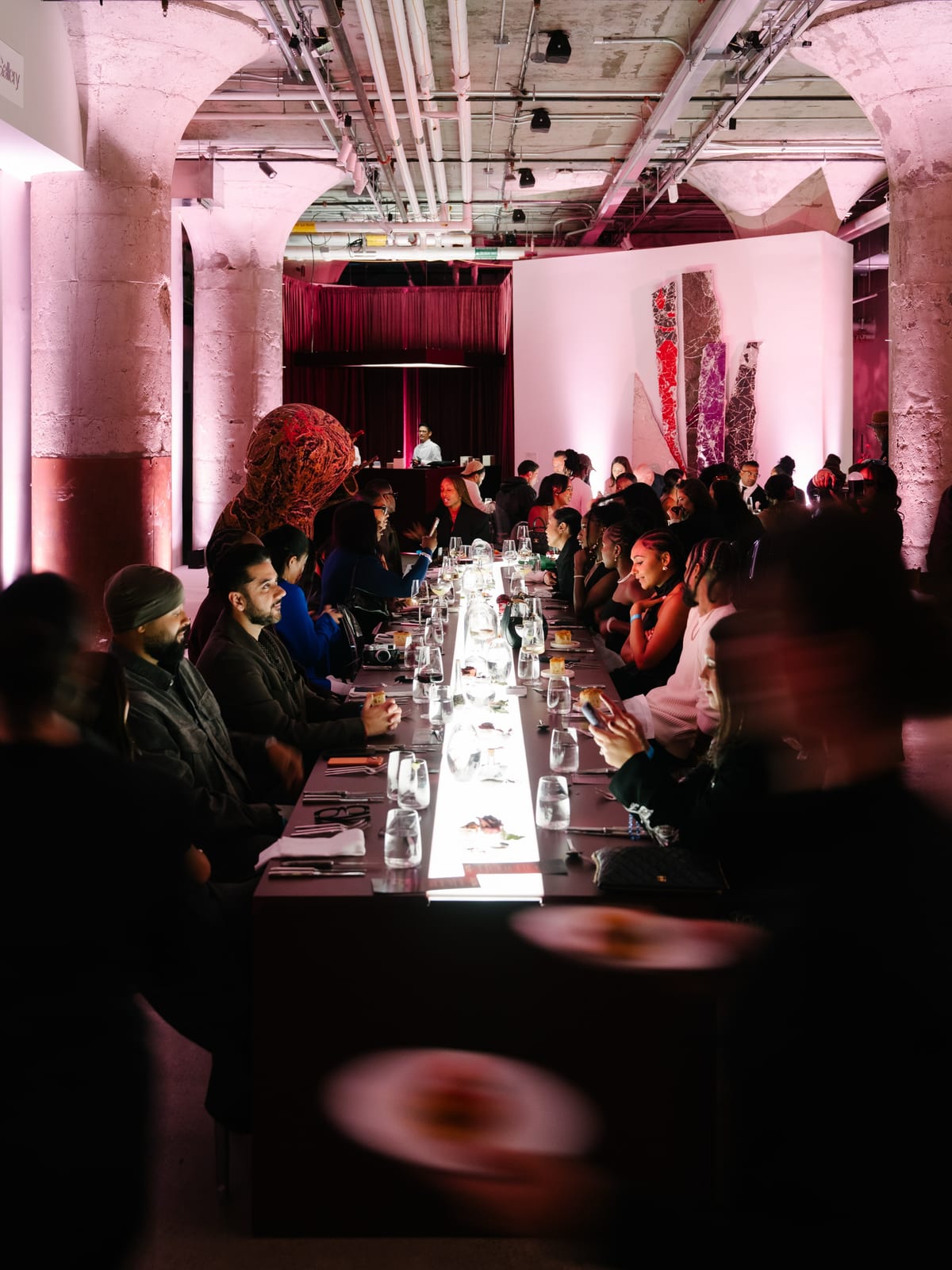 Guests seated along both sides of a long, lit dinner table in a pink-lit room with stone columns and exposed pipes, at the DOSAYDO dinner