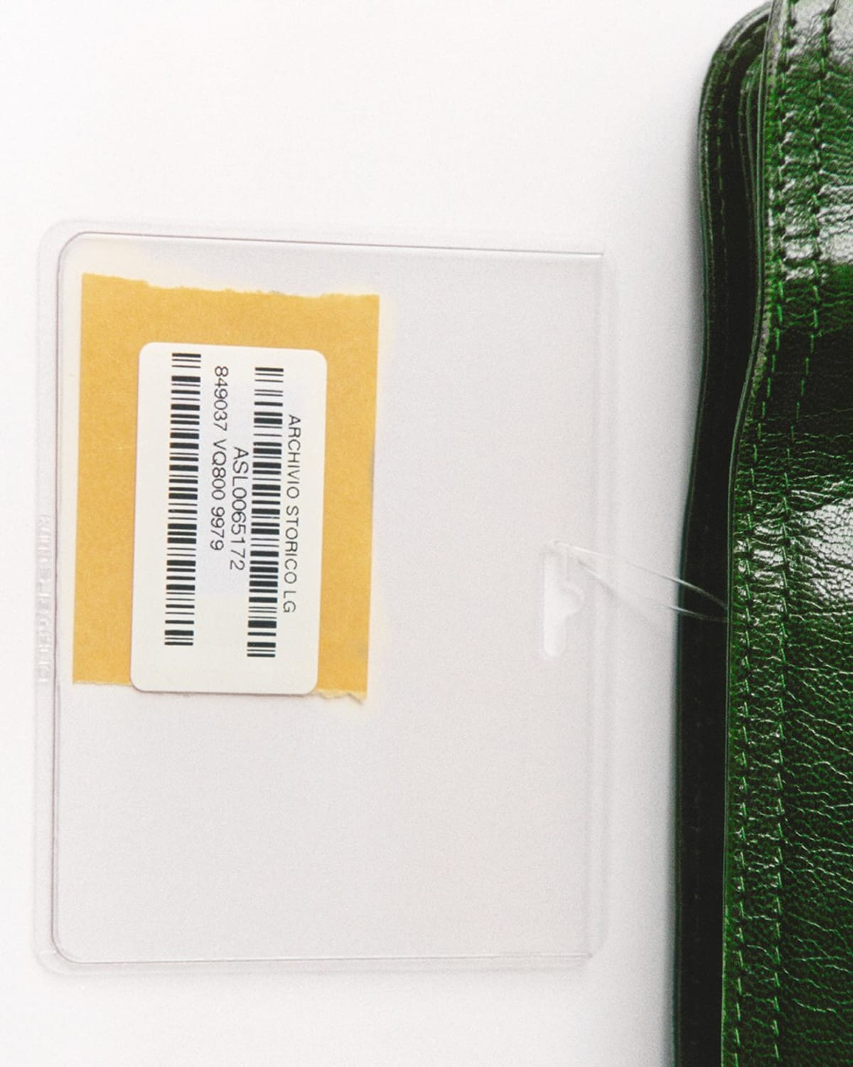 A clear plastic archive sleeve with a barcode label reading Archivio Storico, tagged to a glossy emerald-green leather clutch beside it