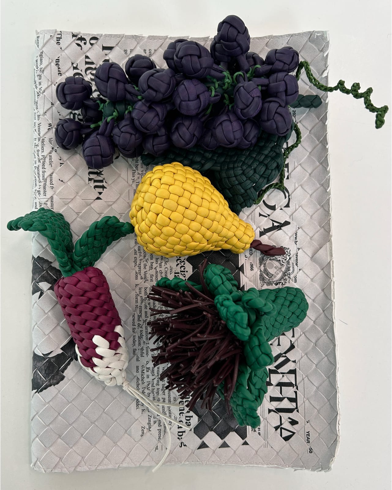 A still life of fruit and vegetables made from woven leather cord, a purple bunch of grapes, a yellow pear, a deep magenta and white radish and a spiky piece of brown cord with green woven leaves, laid on a sheet of woven newspaper print