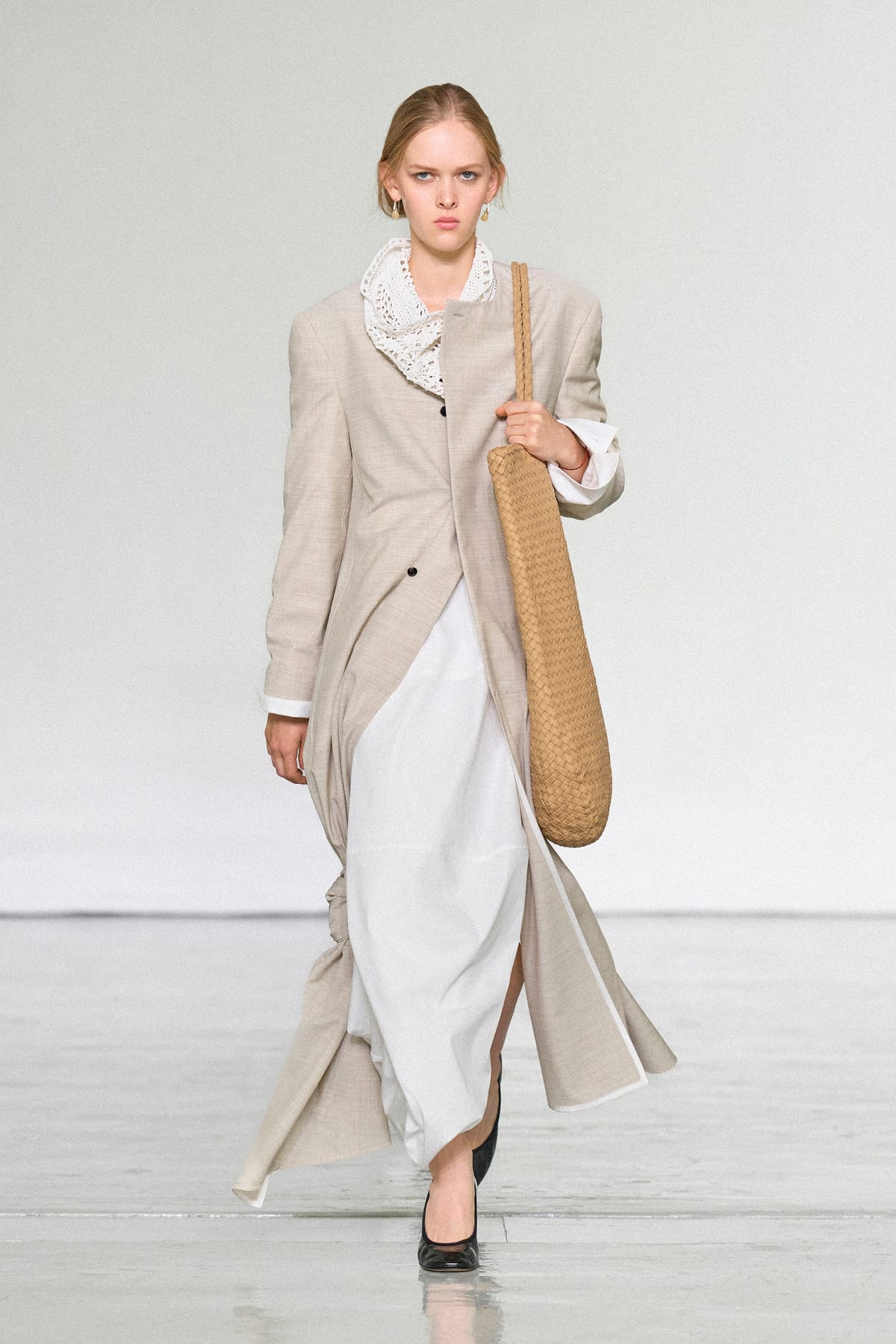 Look 1 of Bottega Veneta's Summer 2027 show: a long beige coat worn open over a white skirt, with a white crochet collar and an oversized tan Intrecciato woven-leather tote on the shoulder