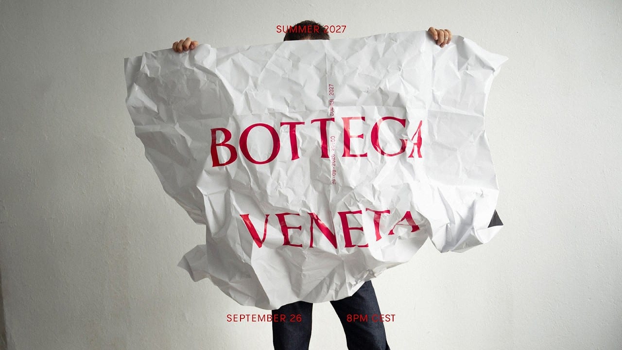 Bottega Veneta's Summer 2027 show invitation: a sheet of crumpled white paper printed with BOTTEGA VENETA in red, held up in front of a white wall, with the date September 26, 8PM CEST