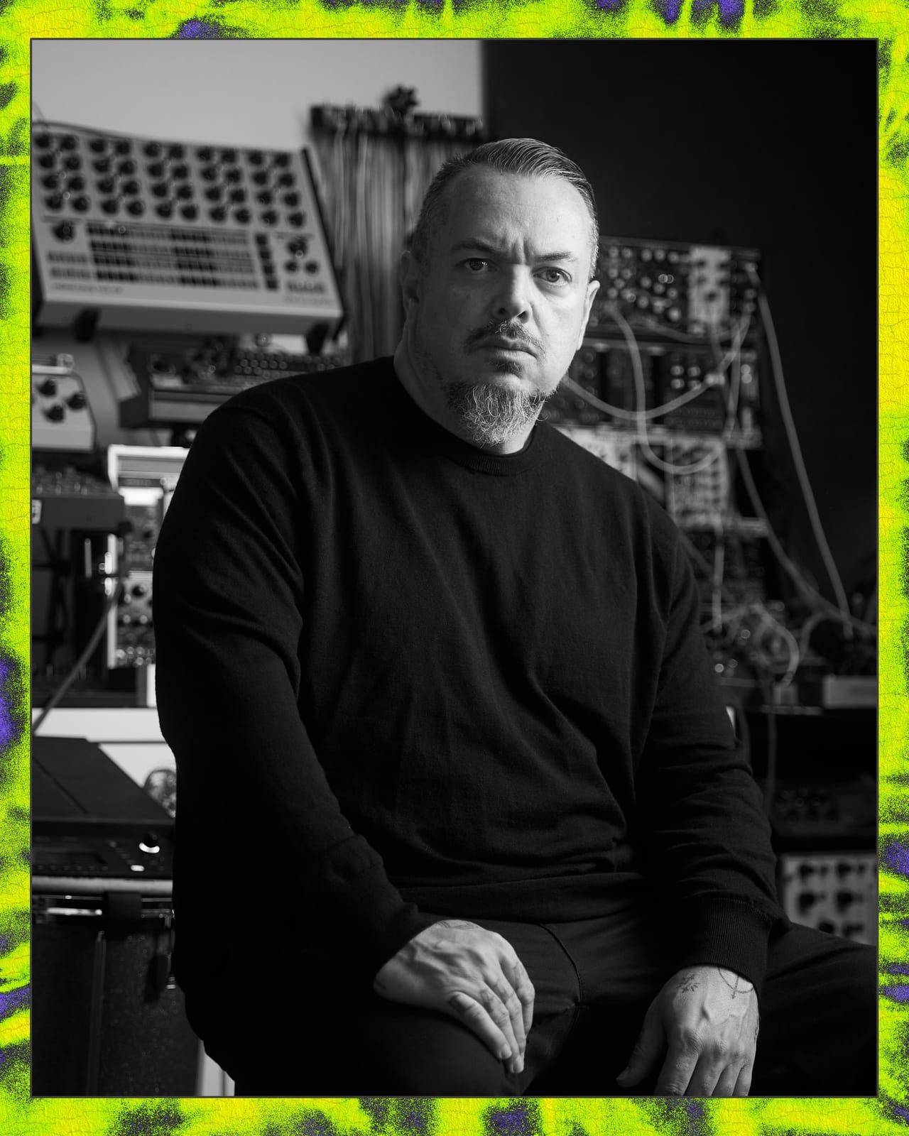 Black-and-white portrait of Iggor Cavalera, with slicked-back hair and a grey goatee, in a dark sweater, seated in front of a wall of modular synthesizers, inside a neon tie-dye border