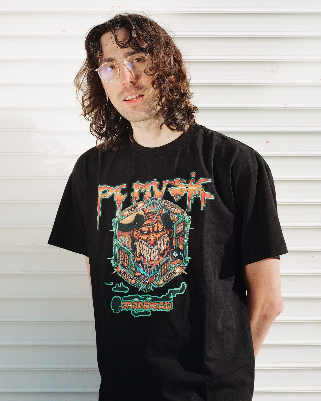 A man with long curly hair and glasses in the black Brain Dead x PC Music POP CRYPT tee, a drippy PC Music logo over a horned creature inside a cube-shaped crest, against a white shutter