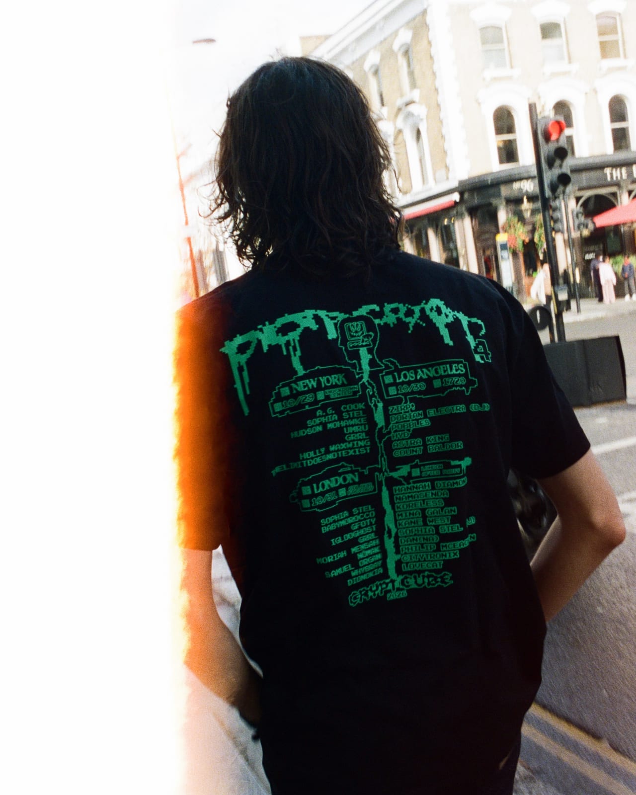 The back of the black POP CRYPT tee, printed in green with the POP CRYPT logo and the New York, Los Angeles and London dates and lineups plus the London after-party, worn outdoors on a city street