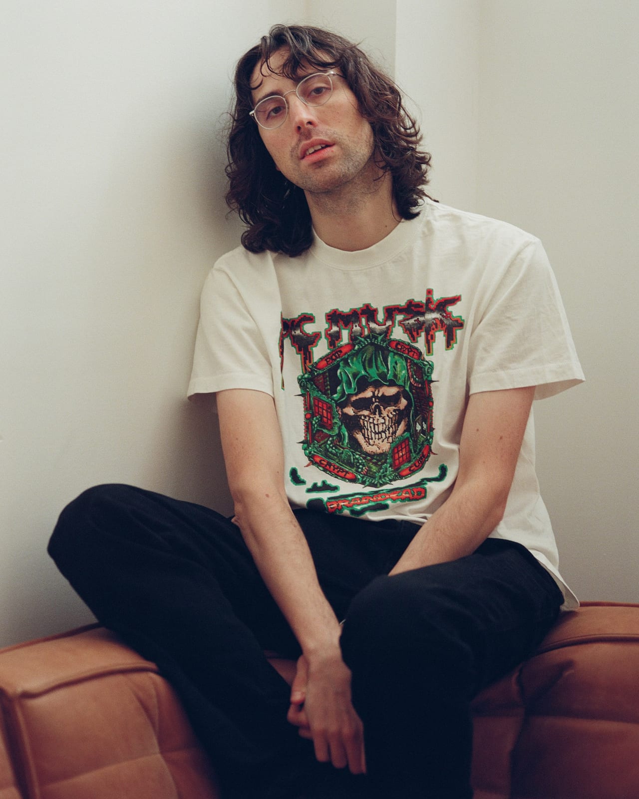 A man with long curly hair sitting on a tan leather bench in the white Brain Dead x PC Music POP CRYPT tee, a hooded skull in green inside the cube crest under the PC Music logo