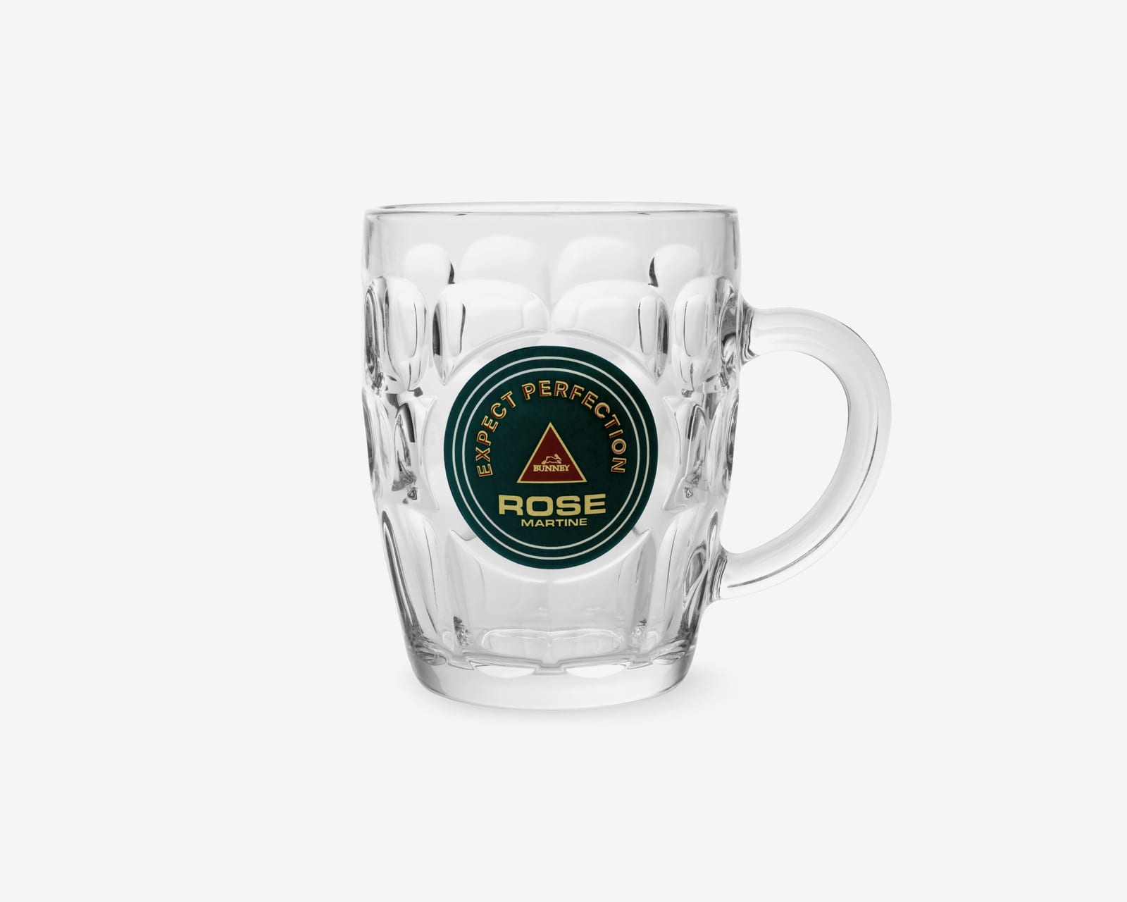 A clear dimpled pint glass with a handle, printed with a round dark green badge reading EXPECT PERFECTION, ROSE, MARTINE around a red triangle