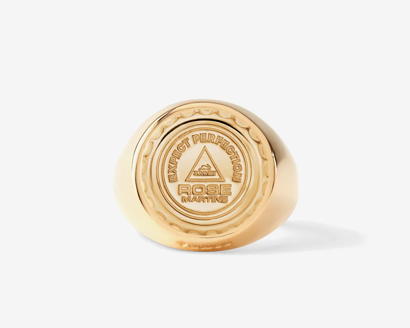 A heavy yellow gold signet ring whose round face is a bottle cap with a crimped rim, embossed EXPECT PERFECTION, a triangle with the Bunney logo, and ROSE MARTINE
